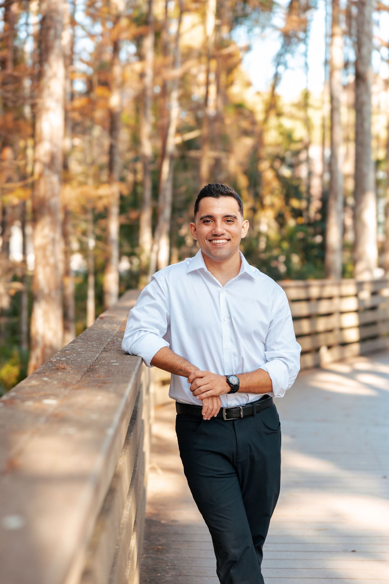 A person in a light blue dress shirt and dark pants stands casually on a wooden boardwalk in an autumn forest setting.