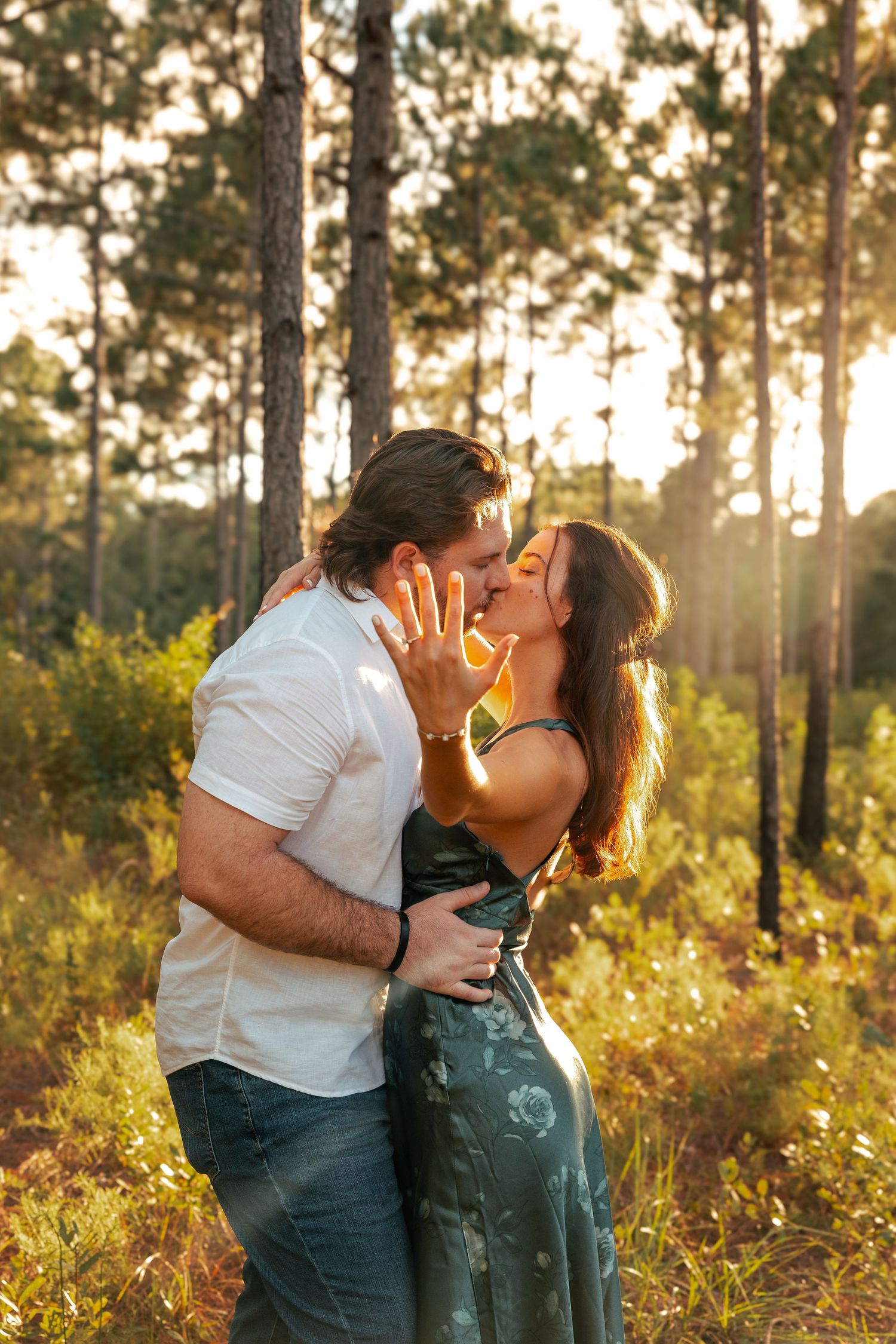 Couple sharing a romantic kiss at golden hour in a sunlit forest, woman showing off engagement ring.