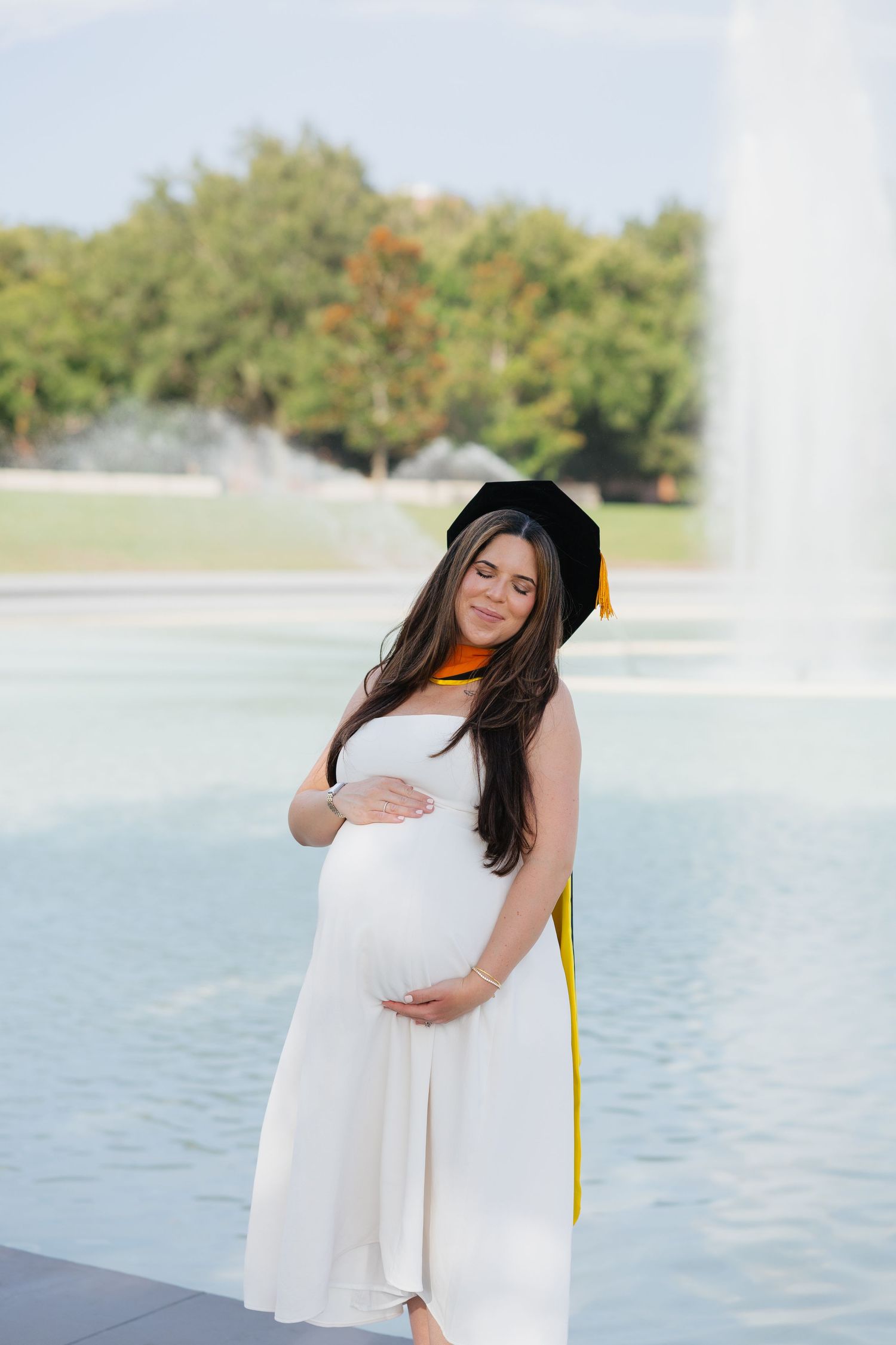 Pregnant woman in white dress and graduation cap stands by fountain, cradling baby bump outdoors.