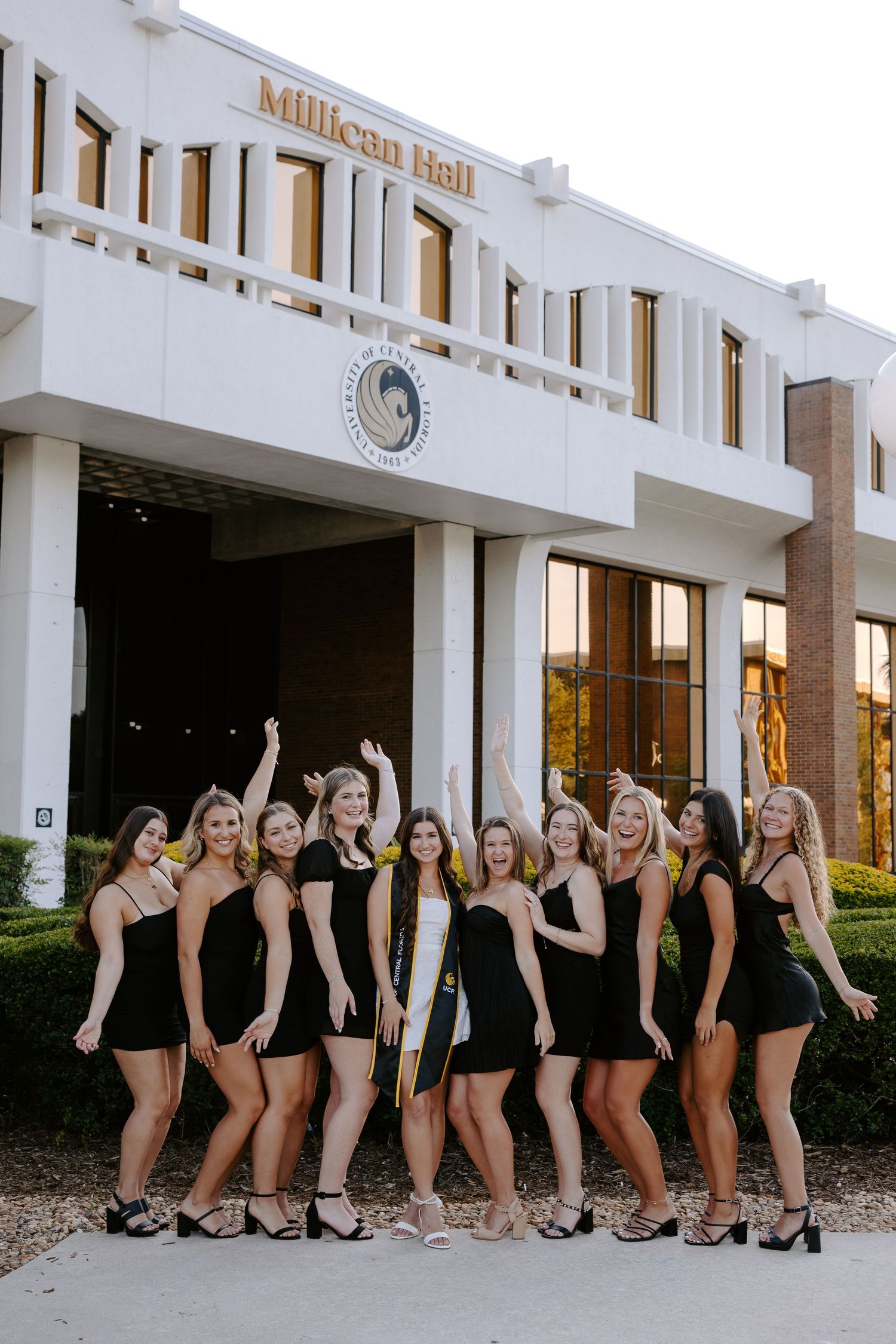 Group of graduates in black attire pose outside white university building in celebratory formation.