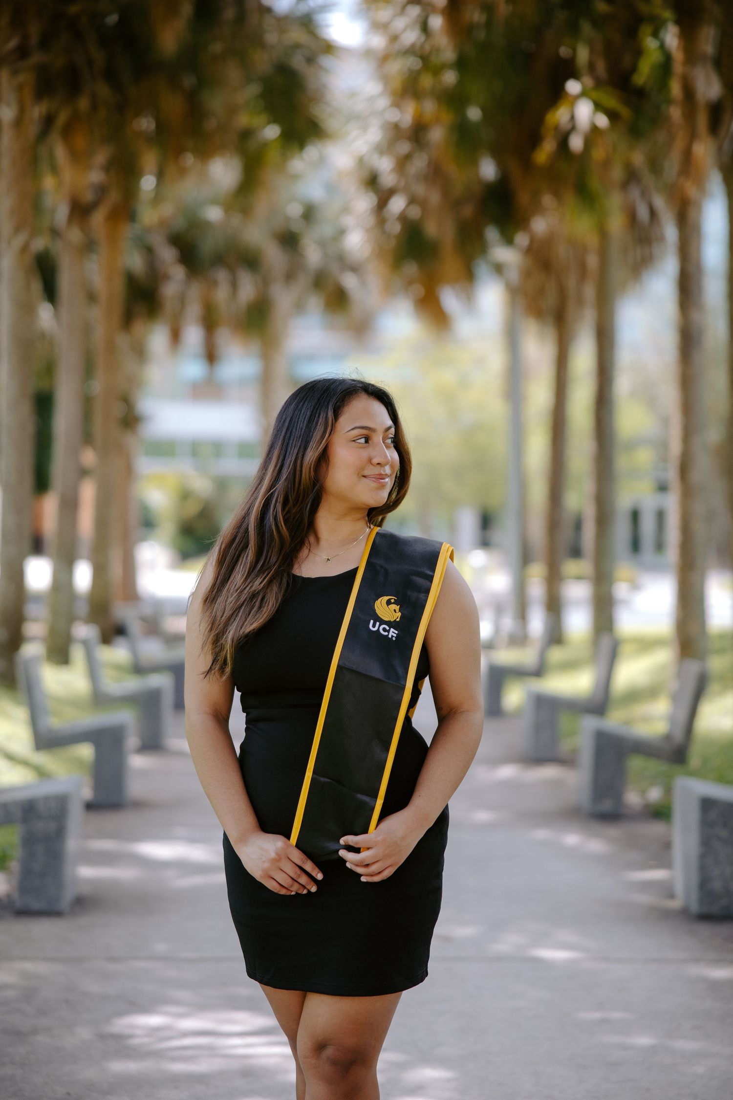 Graduate in black attire poses with UCF stole among palm trees on campus walkway.