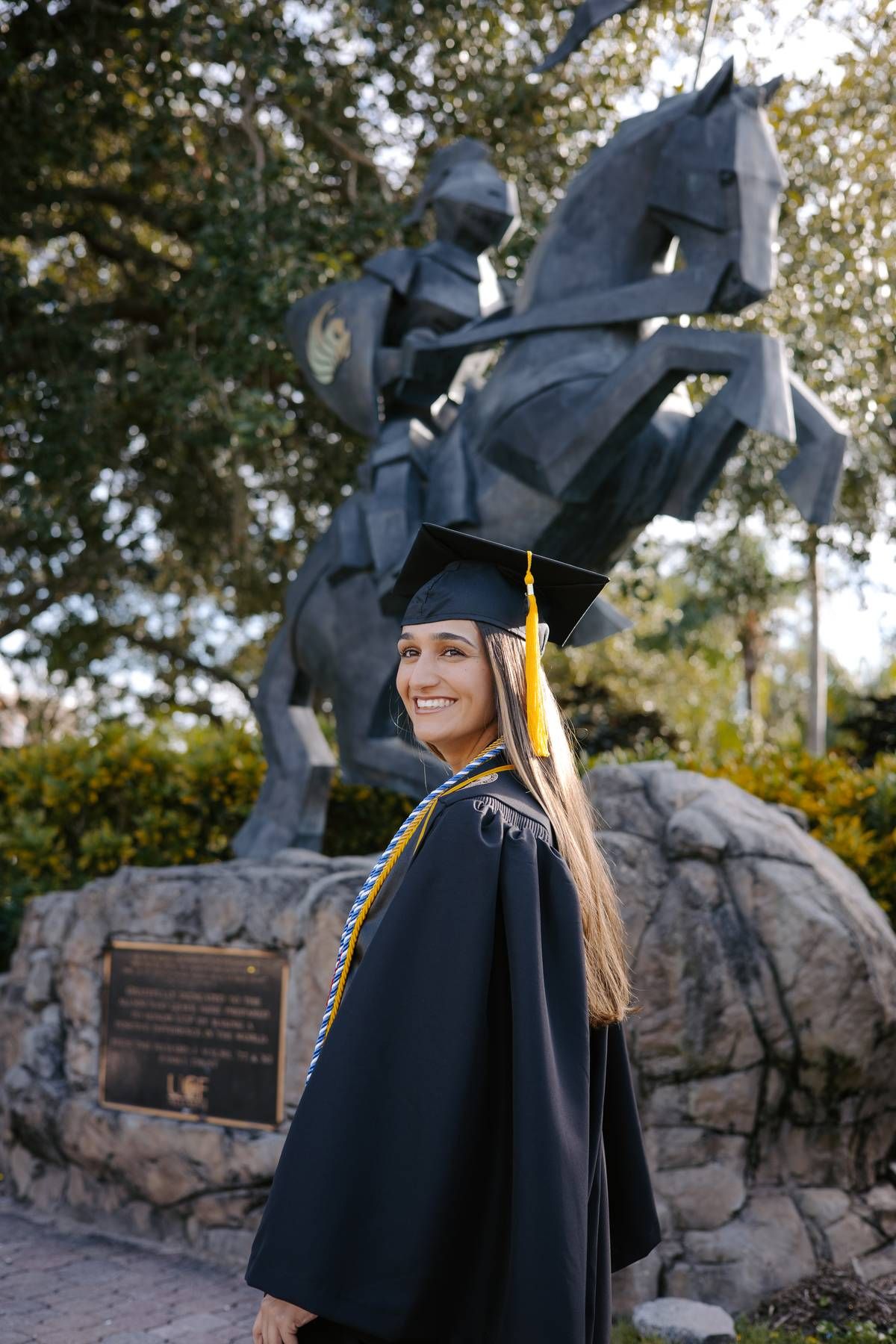 Graduate in traditional black cap and gown posing by outdoor monument.