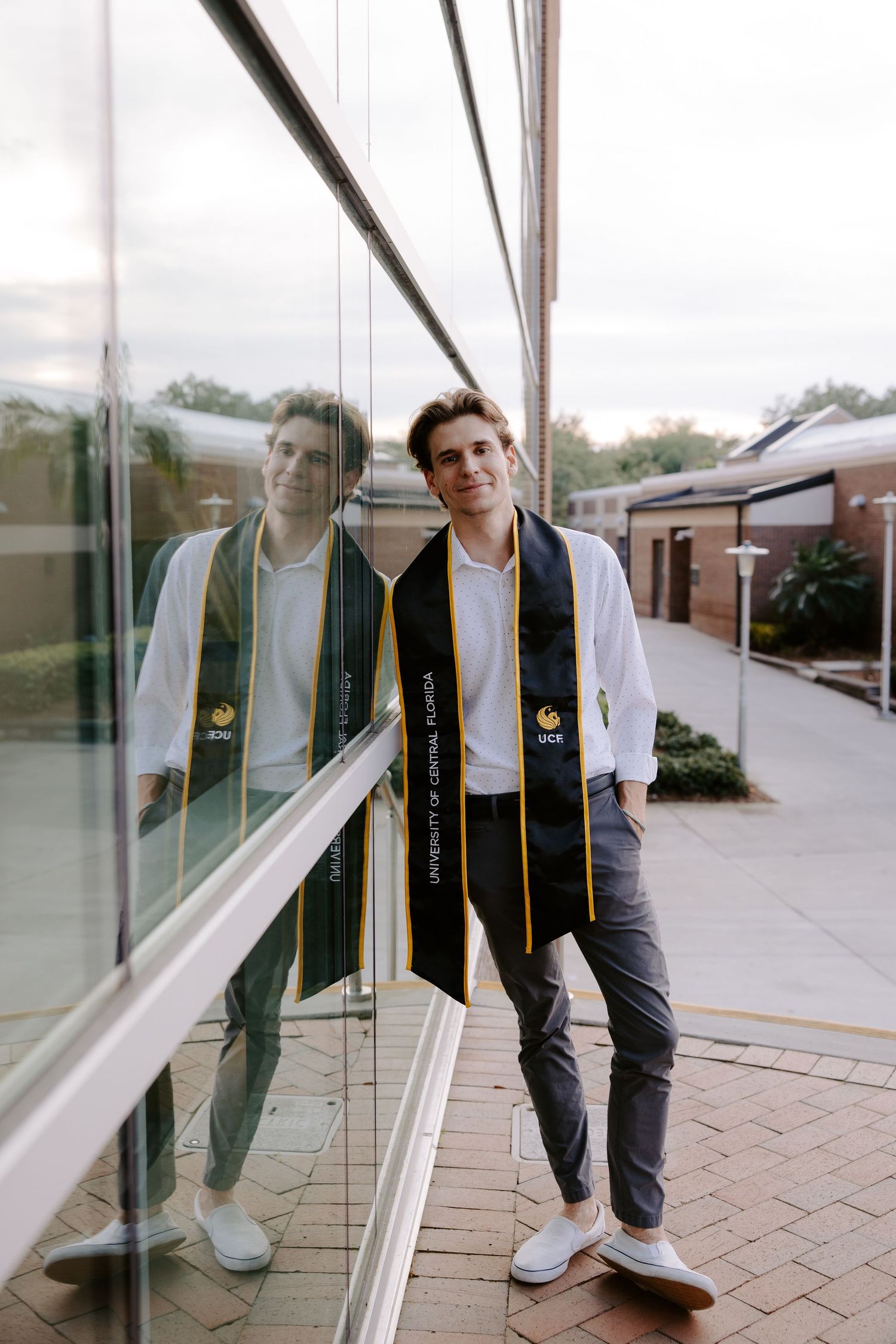 Graduate poses with graduation stole against reflective glass building exterior.