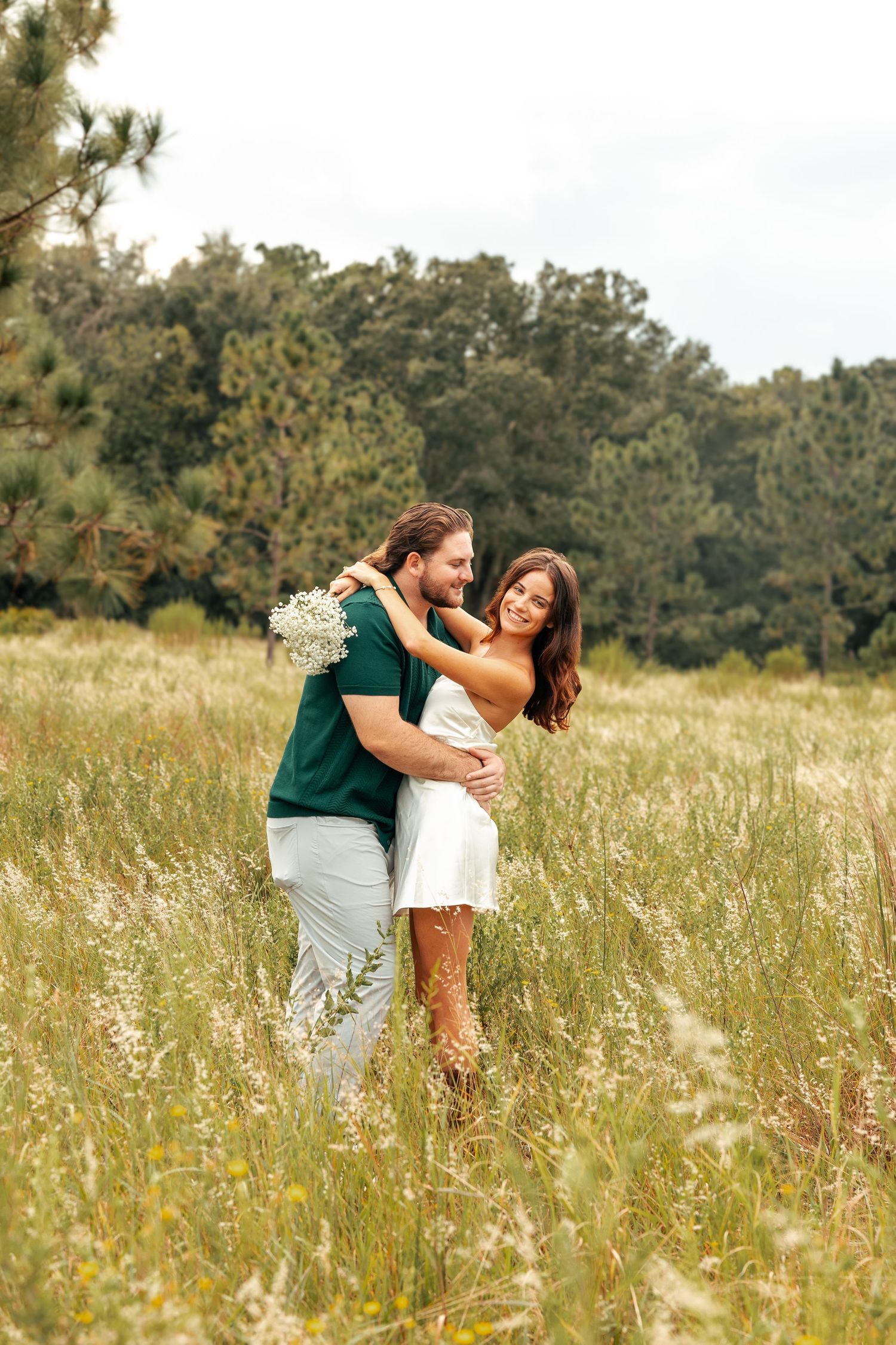A couple embraces in a scenic meadow with tall grass and evergreen trees in the background.