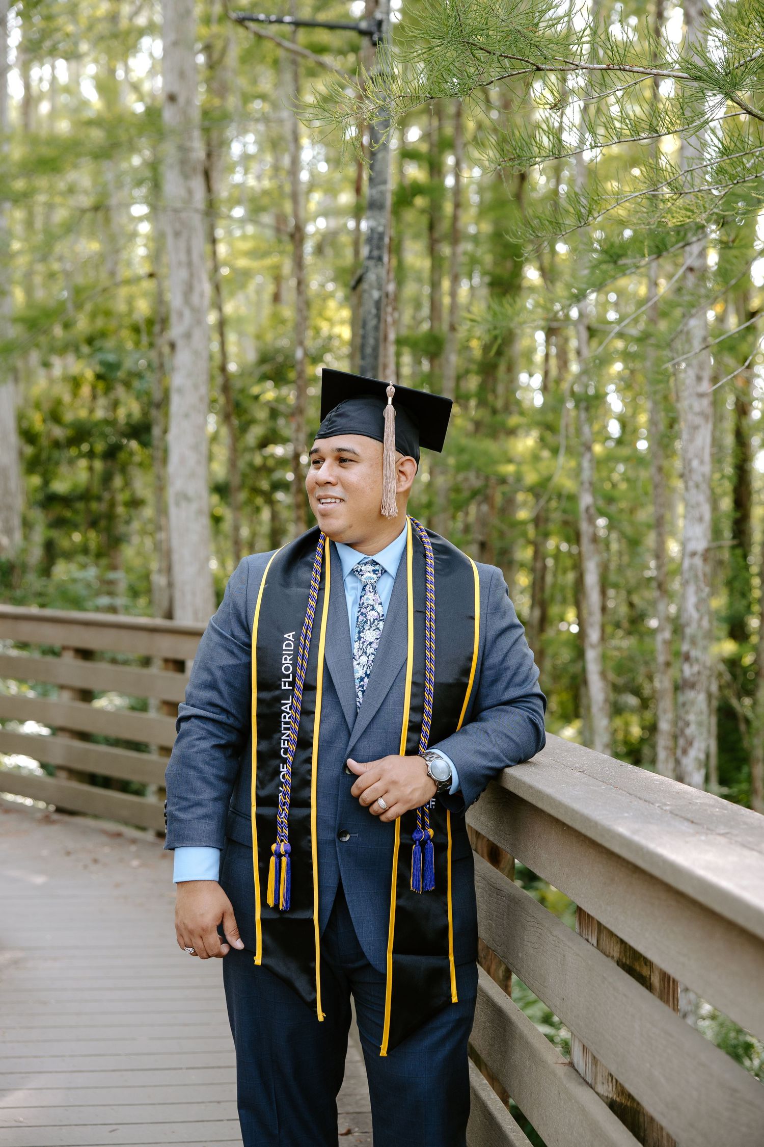 Graduate in blue academic regalia poses on wooden boardwalk surrounded by trees.
