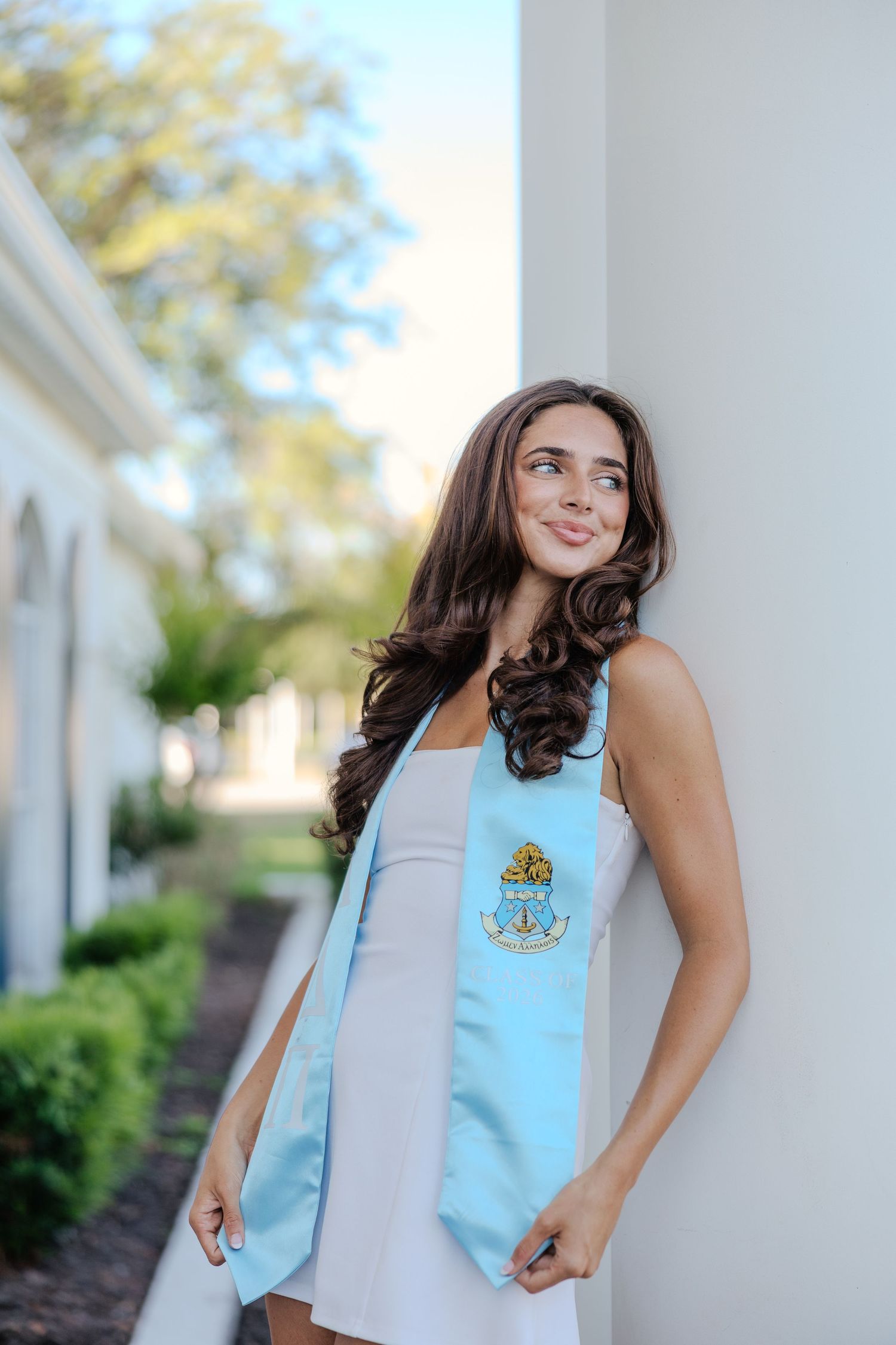 Young woman in white dress wearing light blue graduation stole with emblem, leaning against white column outdoors.