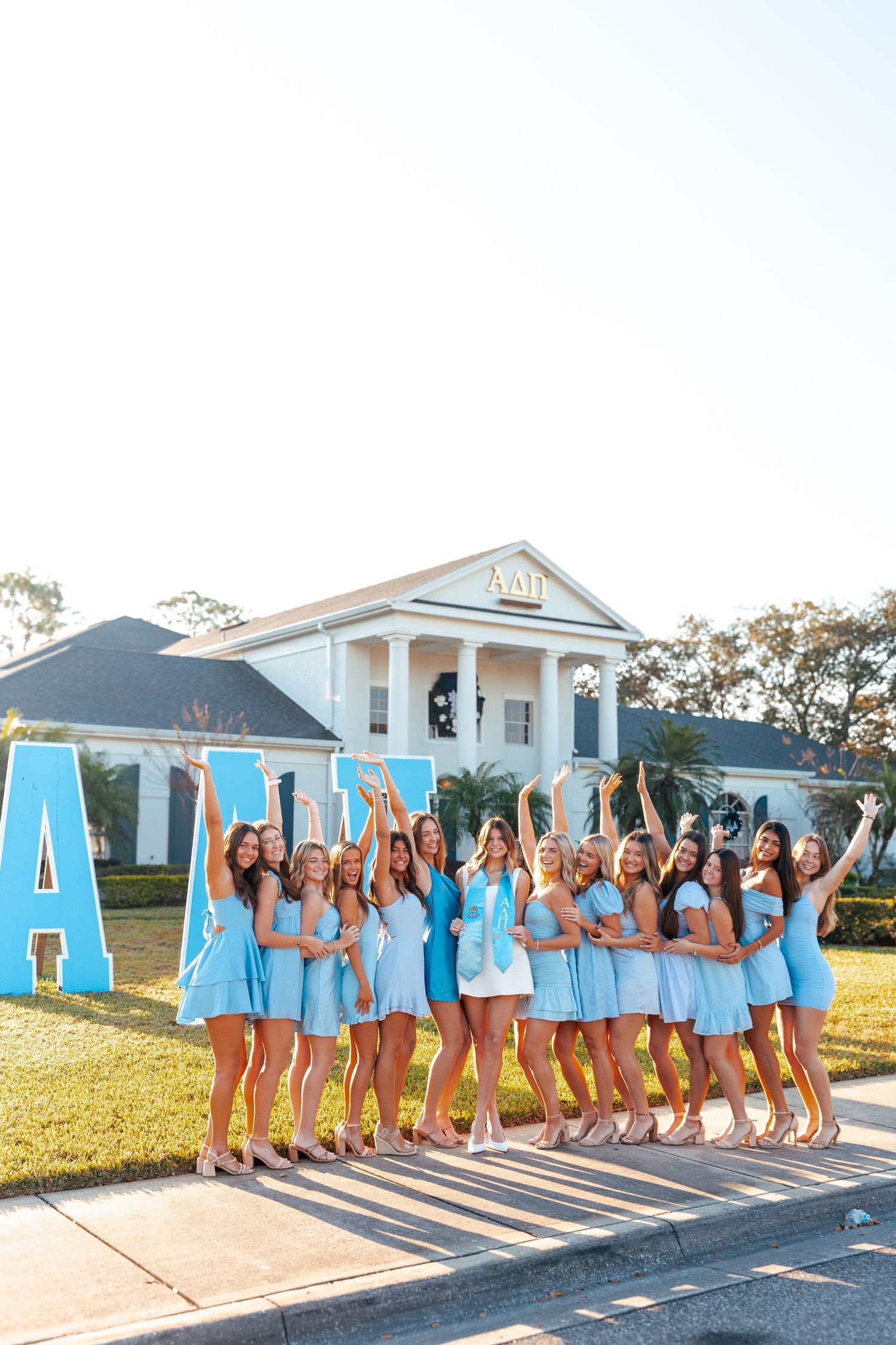 Group of sorority women in light blue dresses posing in front of Alpha Delta Pi house with large Greek letters.