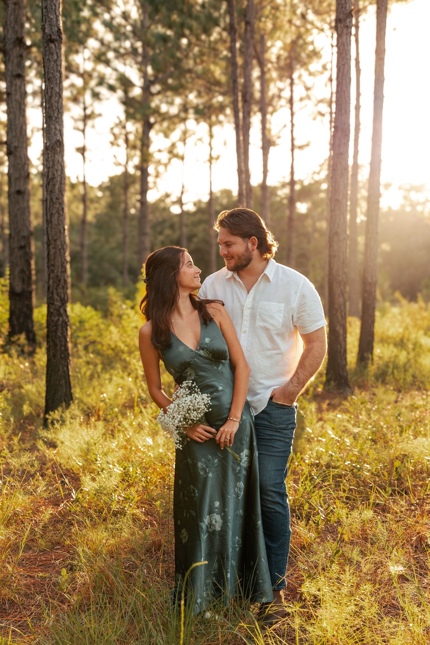 A couple shares an intimate moment in a sunlit pine forest during golden hour, surrounded by tall trees and grass.