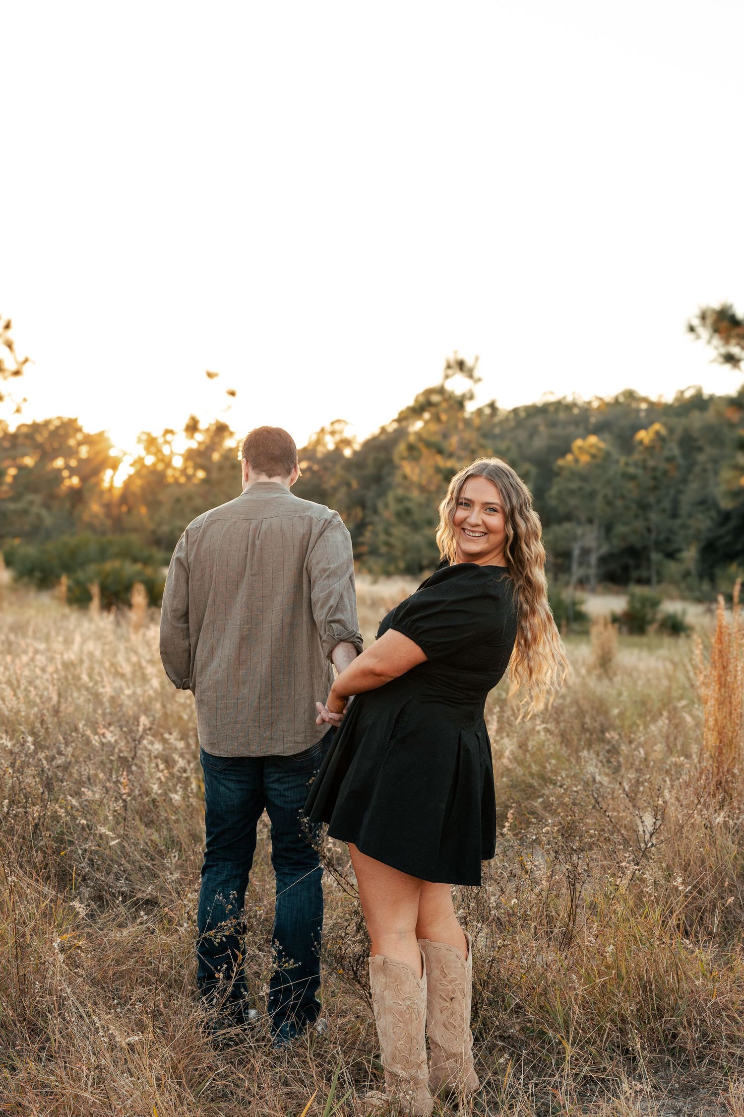 A couple walks through a golden field at sunset holding hands in a romantic outdoor setting.