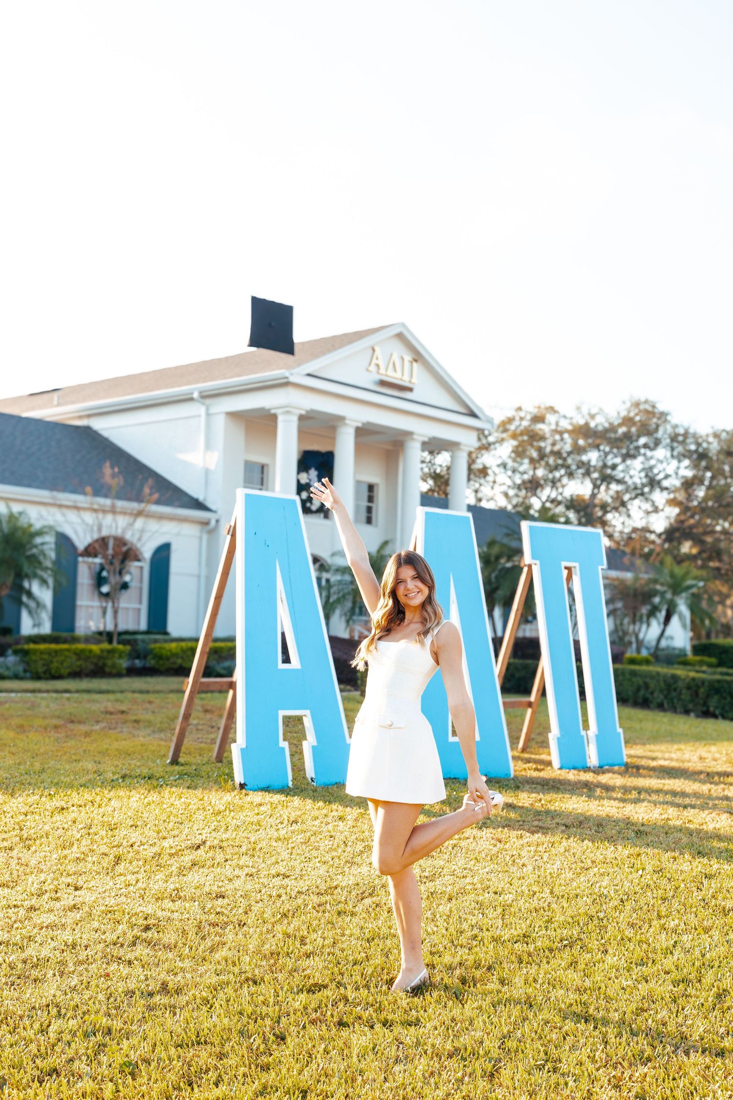 Person in a white dress poses playfully with large blue letters spelling 'ALOHA' on a sunny lawn near white buildings.