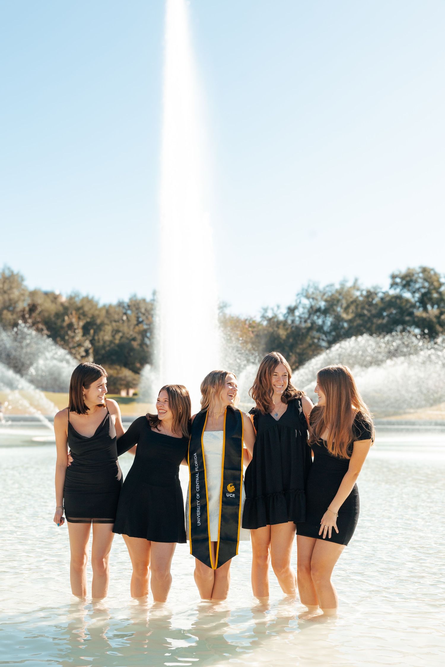 A group of friends in matching black outfits pose together while standing in shallow water on a sunny day.
