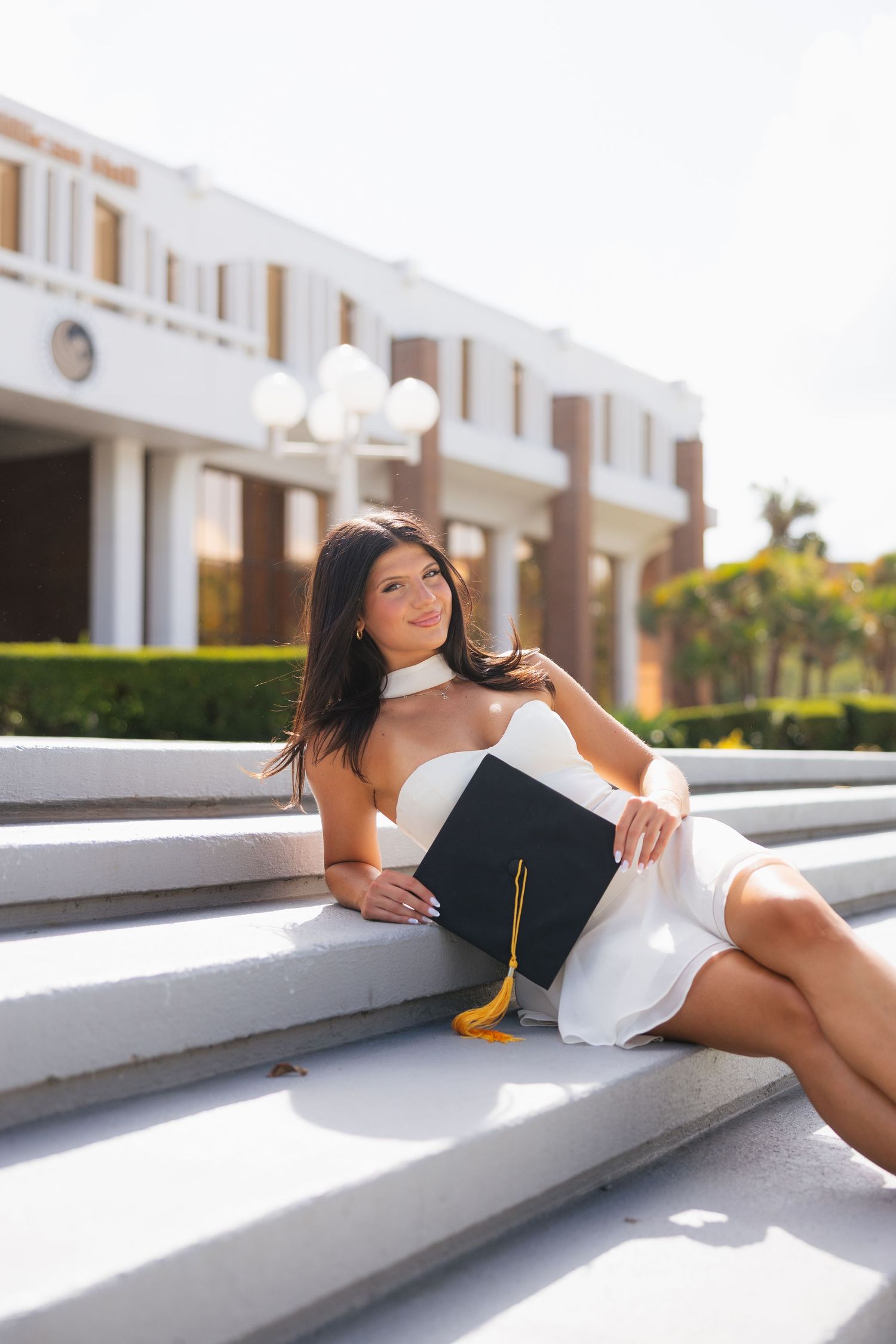 Young woman in white dress holds graduation cap while sitting on steps outside a university building.