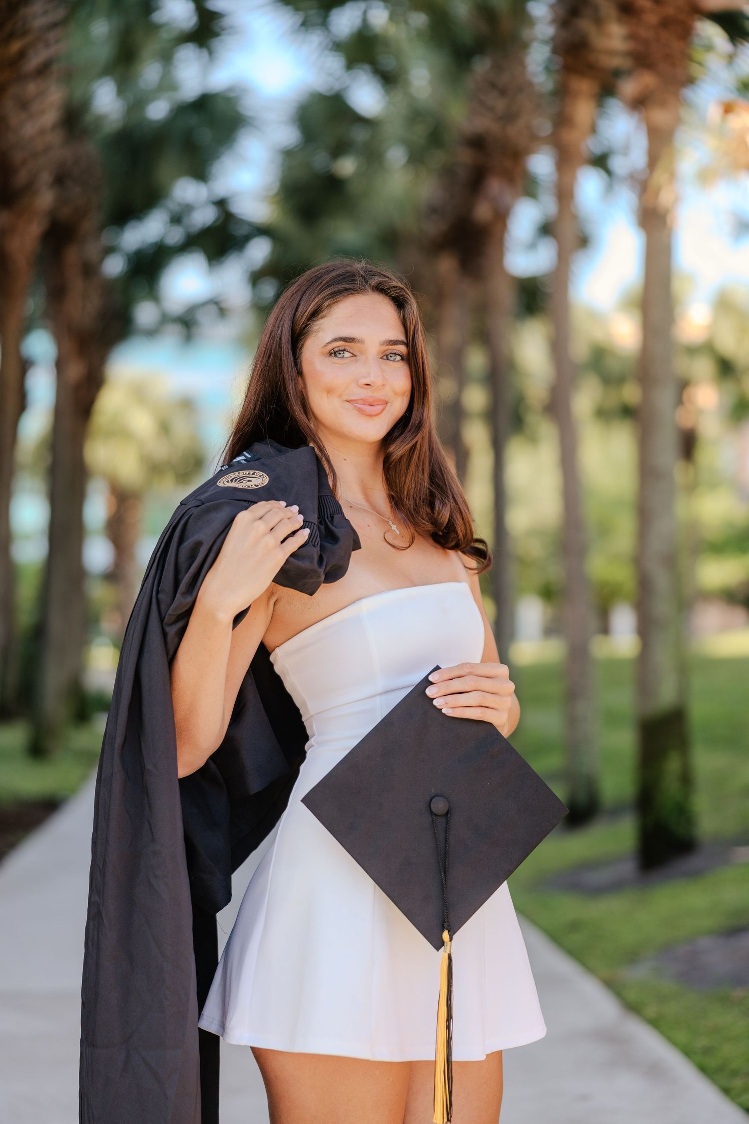 Young woman in white dress holding graduation cap and gown, smiling outdoors among palm trees on a sunny day.