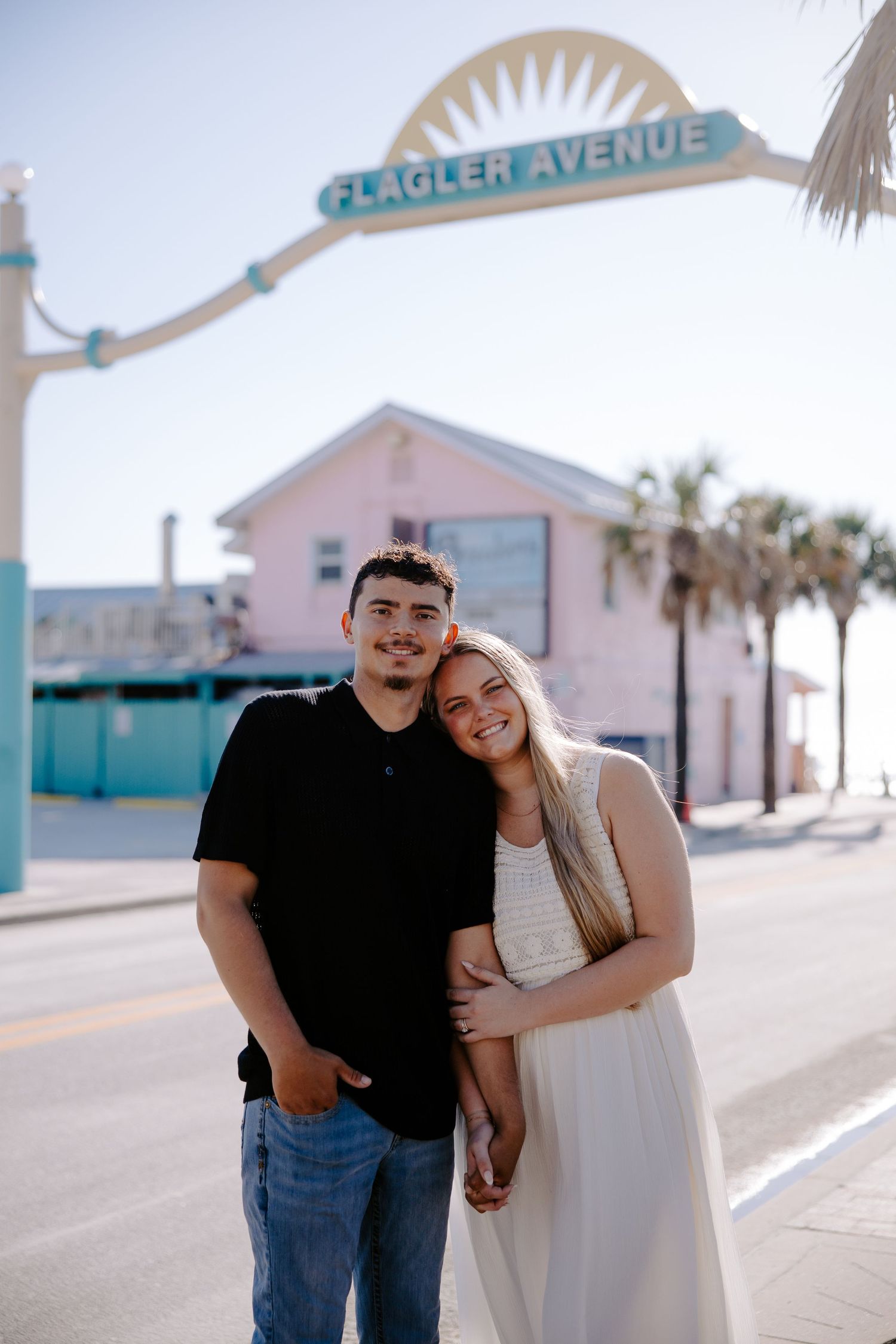 A couple poses together on a sunny boardwalk with palm trees and pastel buildings in the background.