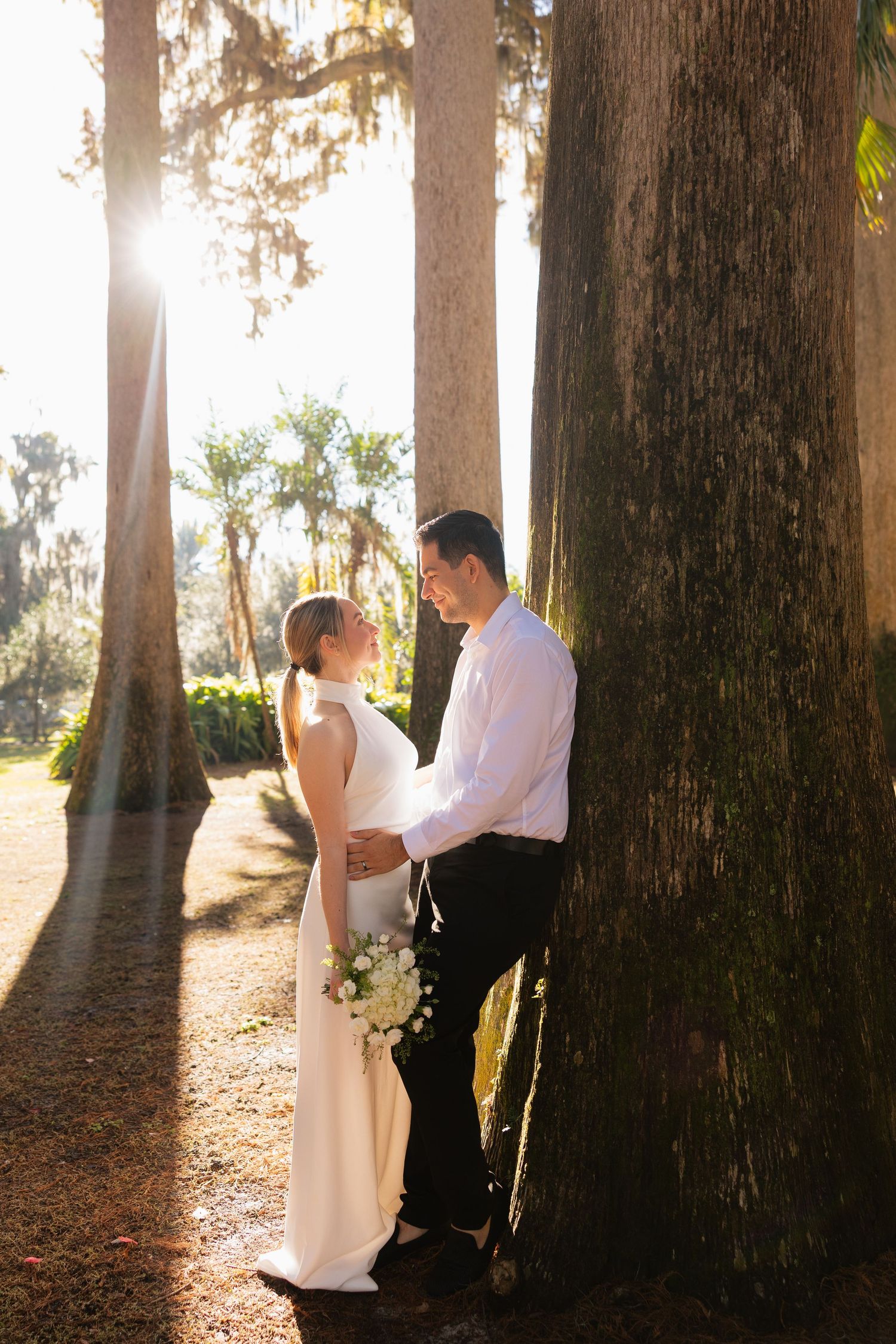 Bride and groom share a romantic moment among tall trees with golden sunlight streaming through in an outdoor wedding portrait.