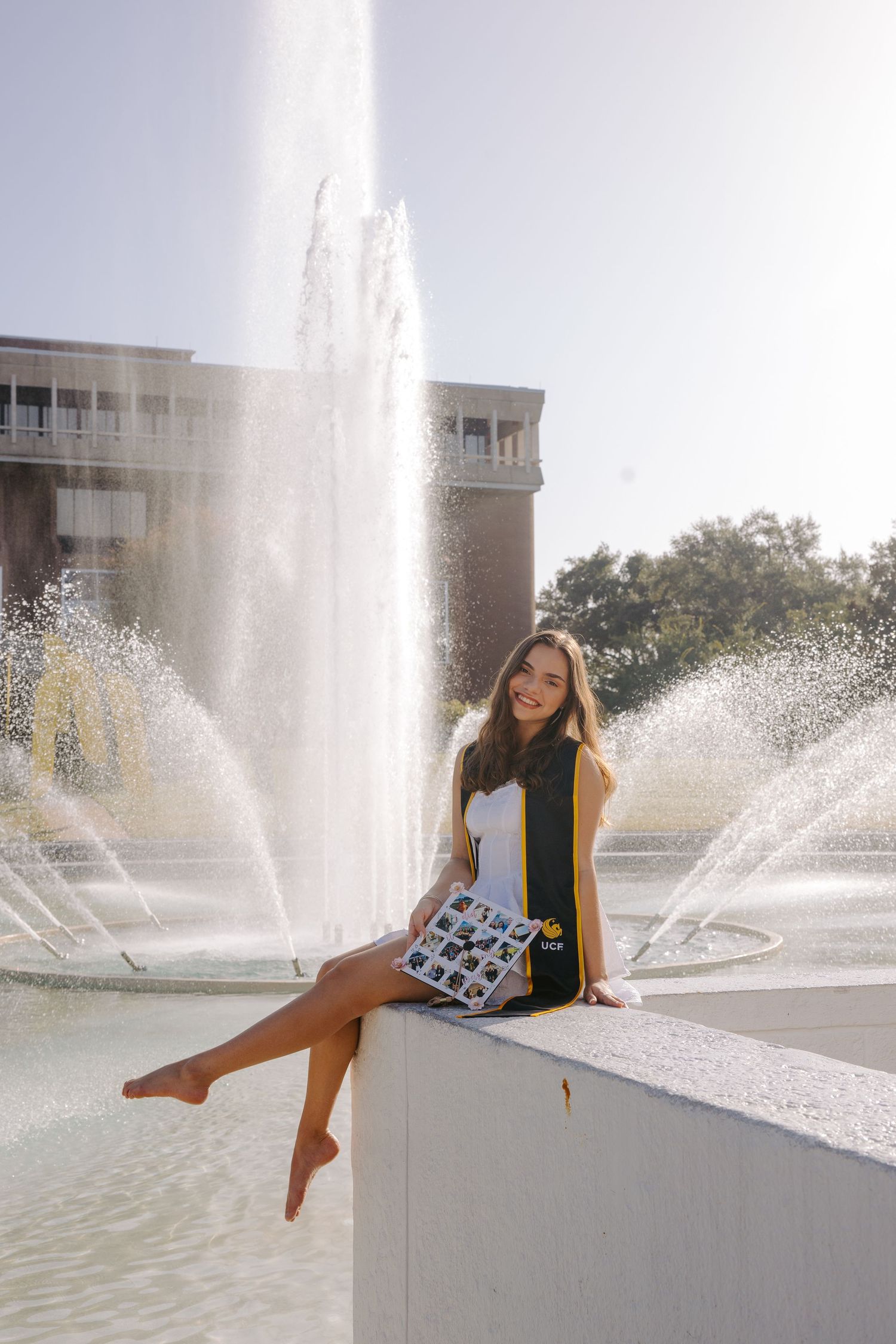 Graduate poses playfully by campus fountain during sunny graduation photo session.