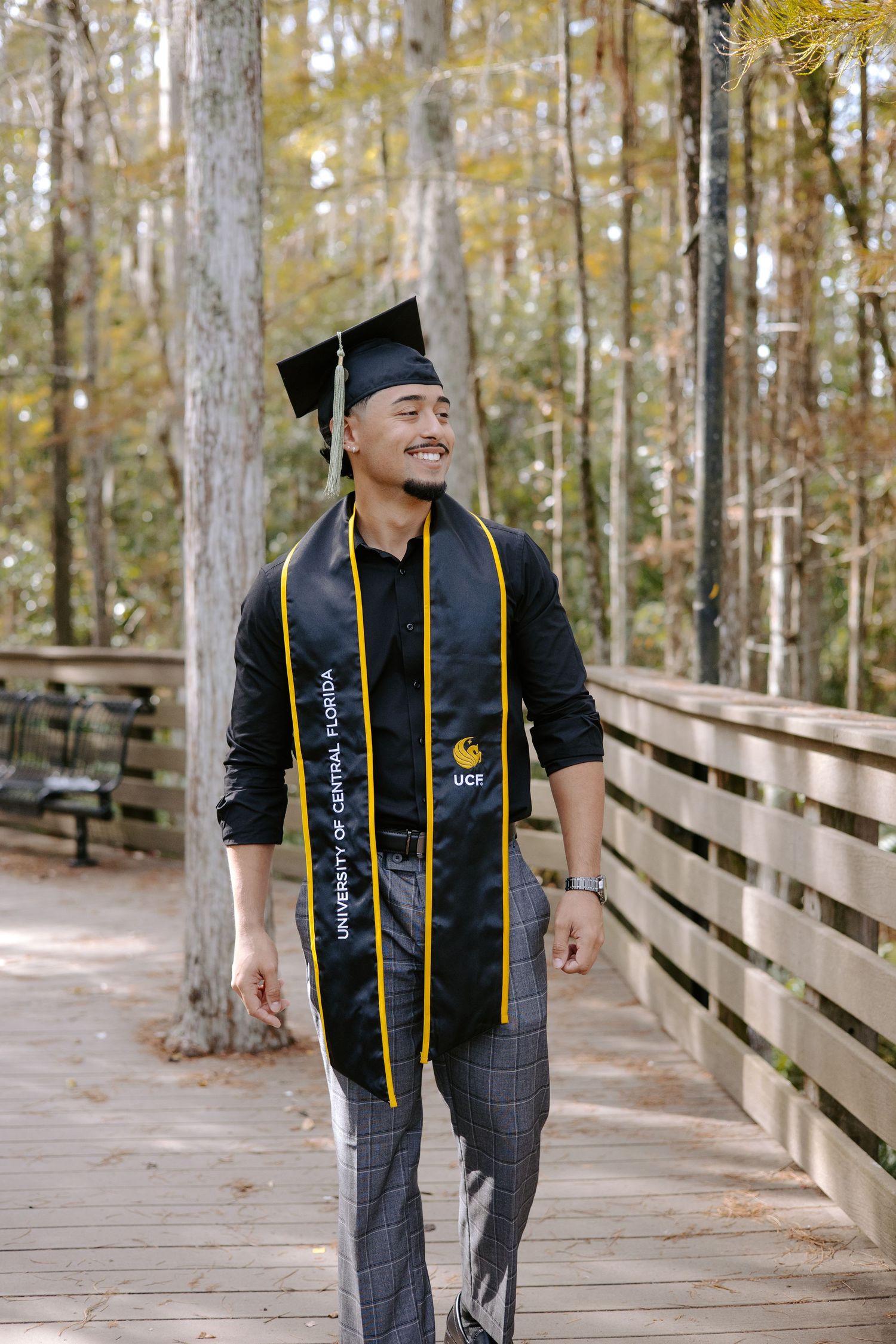 Graduate in cap and gown poses on a wooden boardwalk surrounded by trees during graduation photoshoot.