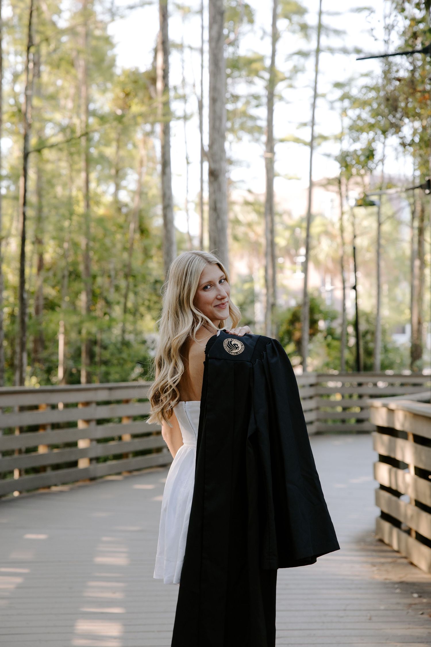 A person poses with a graduation robe outdoors on a wooden boardwalk surrounded by trees.