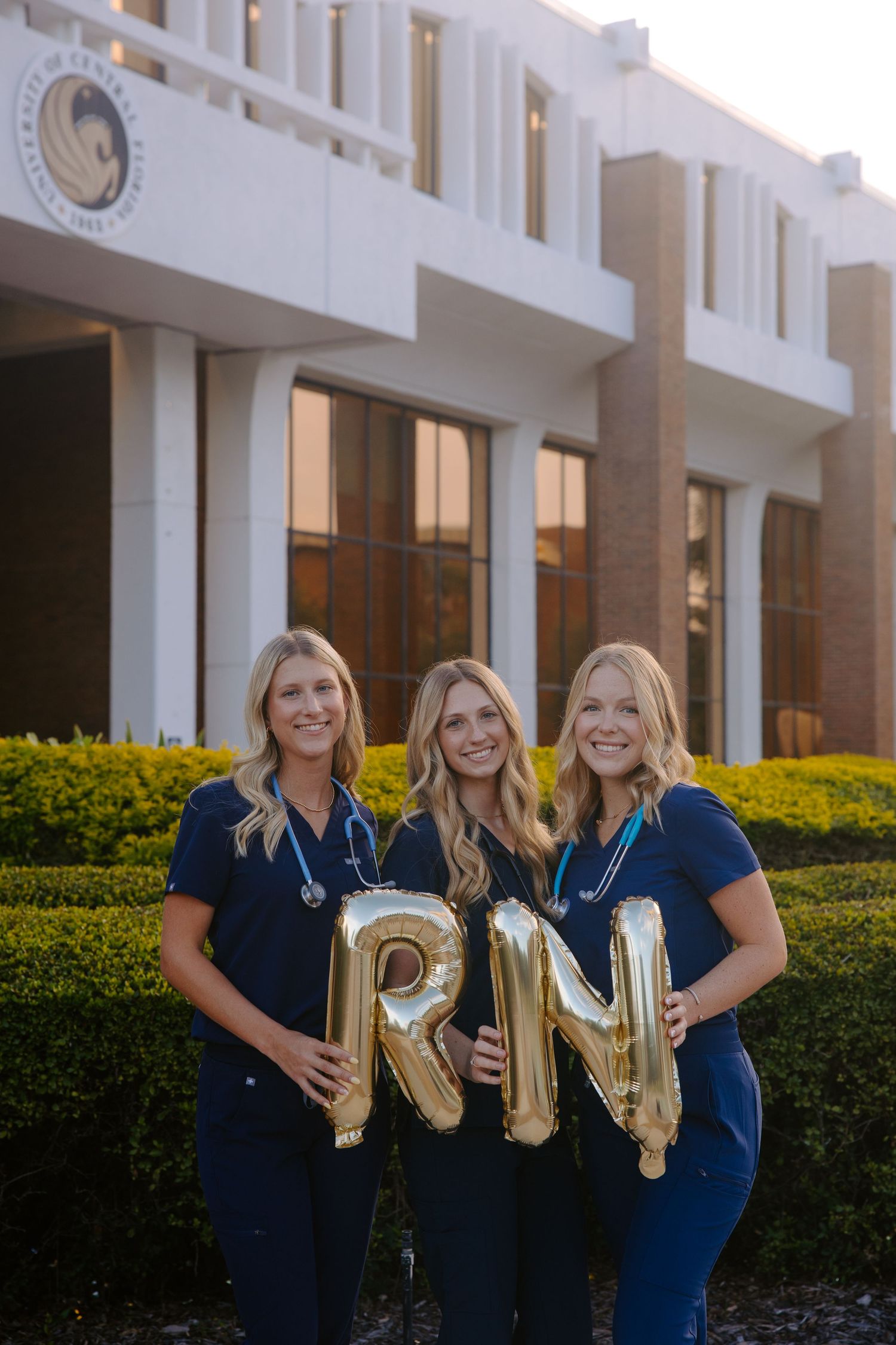 Nursing students pose with gold RN letter balloons outside a medical building.
