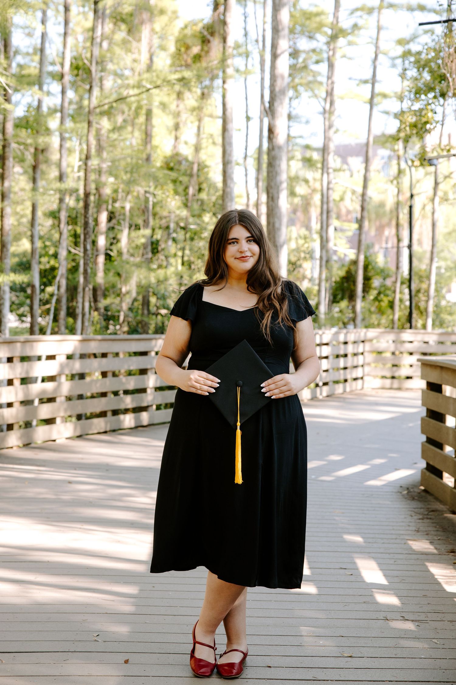 Graduate in black dress poses on wooden boardwalk surrounded by pine trees.