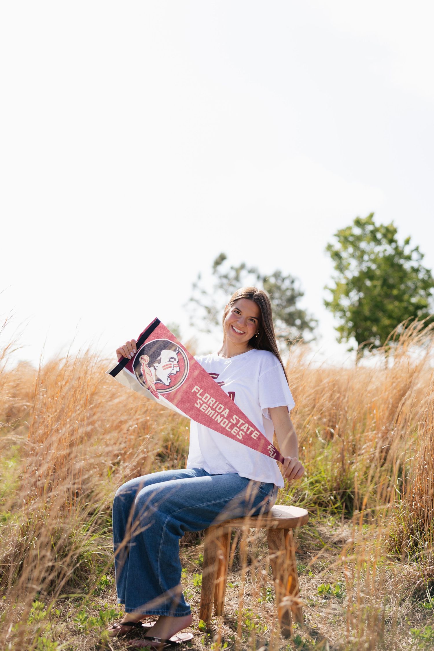 Smiling young woman sitting outdoors holding a Florida State Seminoles pennant flag in a golden grass field.