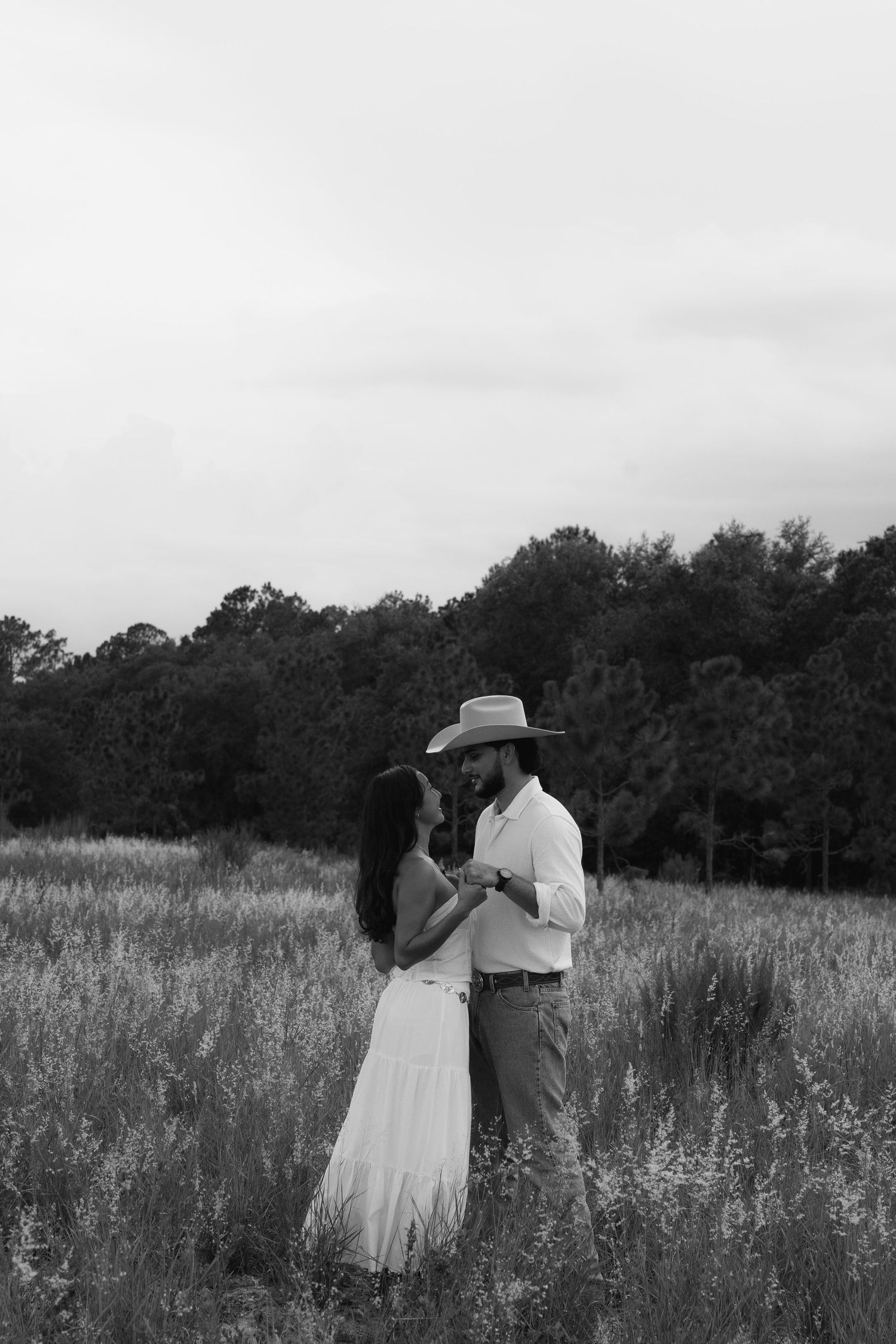 A couple embraces in a dreamy black and white meadow scene with tall grass and trees in the background.