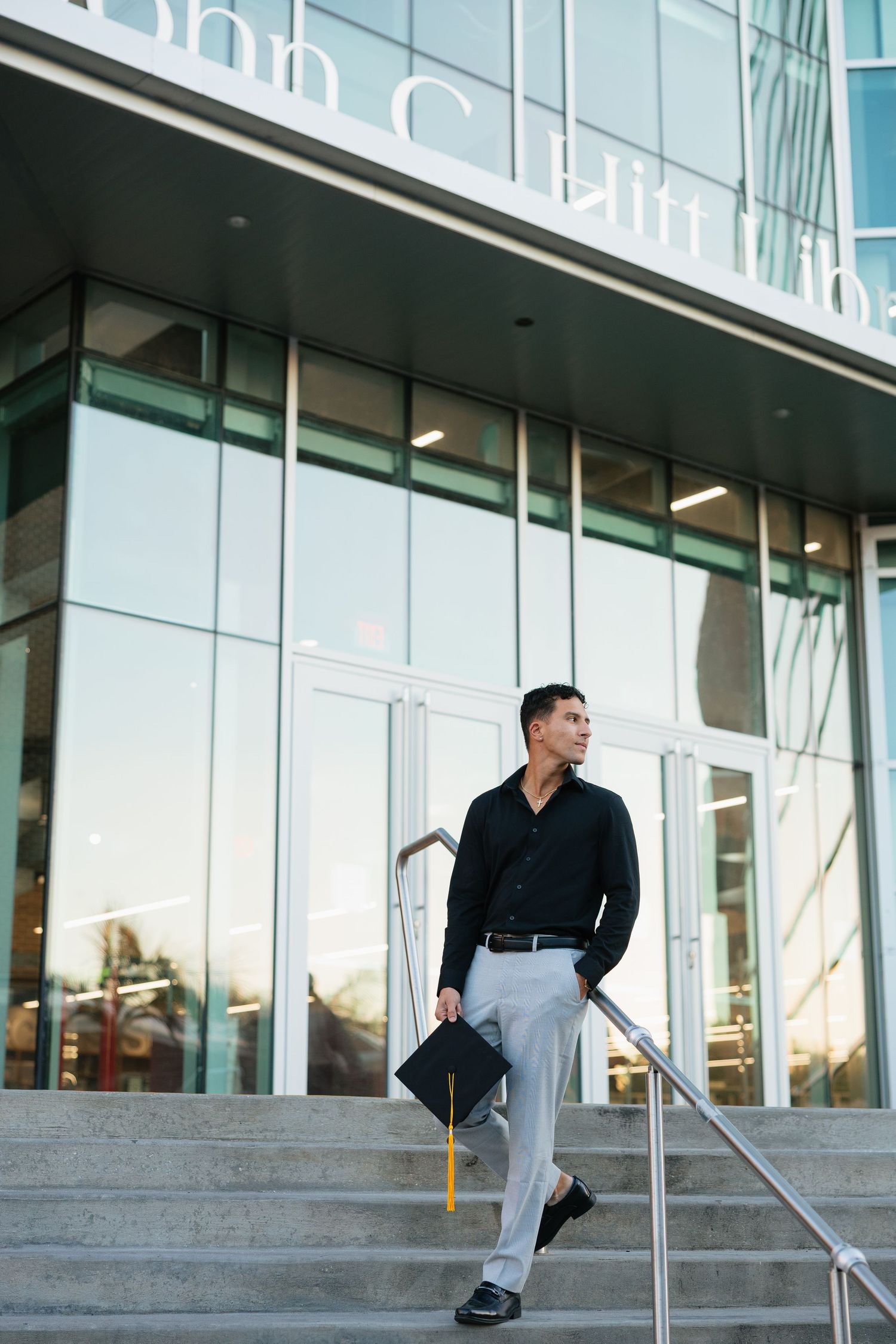 Graduate in business attire stands on steps outside modern glass building holding diploma at sunset.