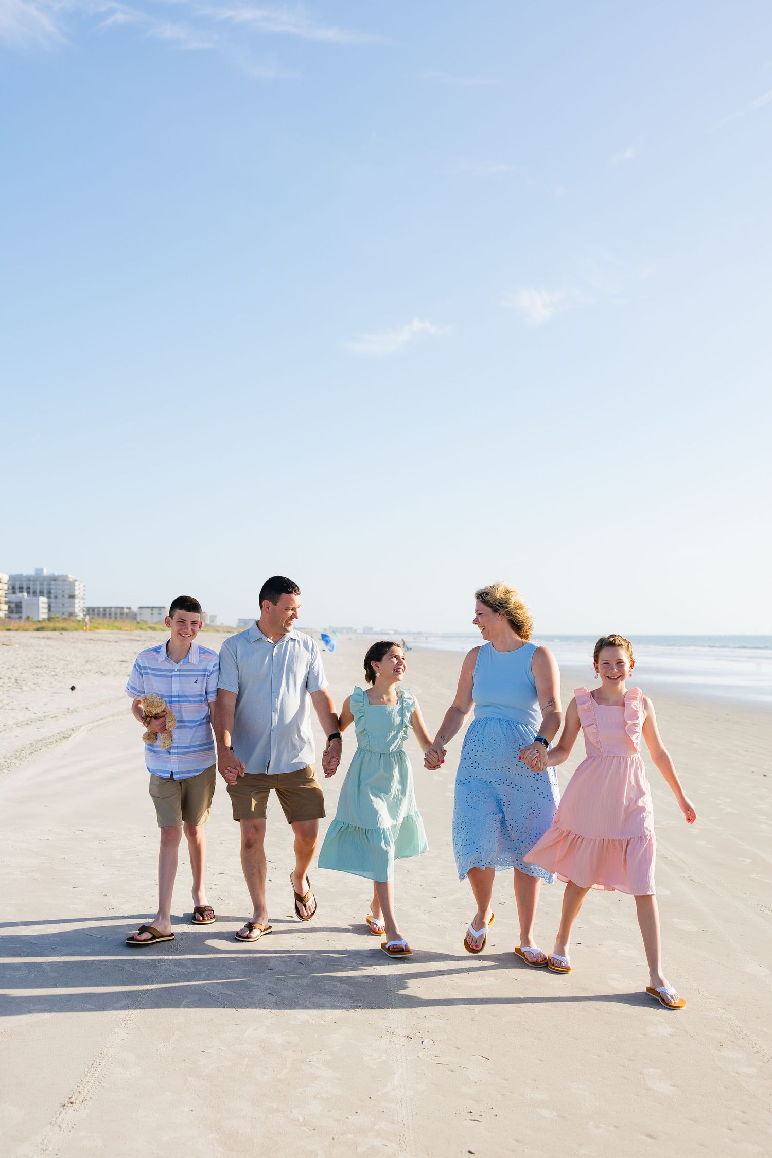A happy family of five walks hand-in-hand along a sunny beach, wearing casual summer outfits in blue and pink.