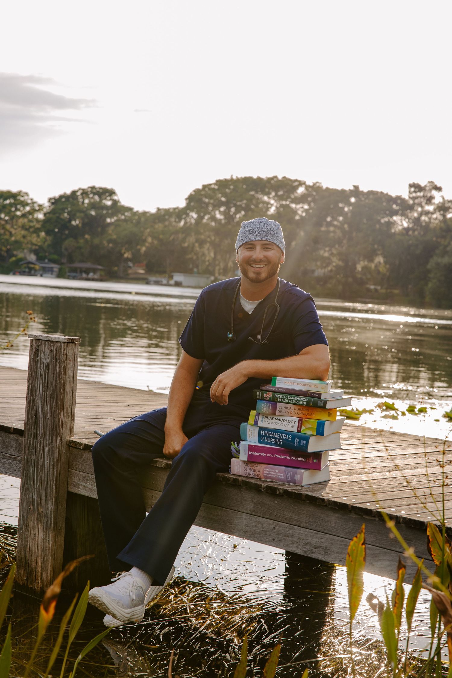 Healthcare worker sits on dock with textbooks beside lake during sunset.