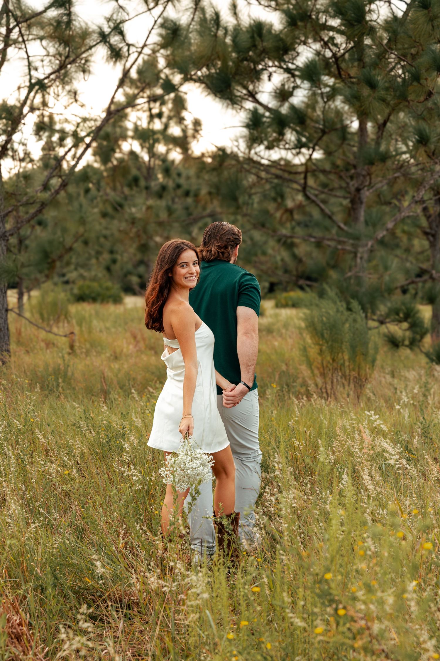 Romantic couple embraces while standing in a golden meadow surrounded by evergreen trees.