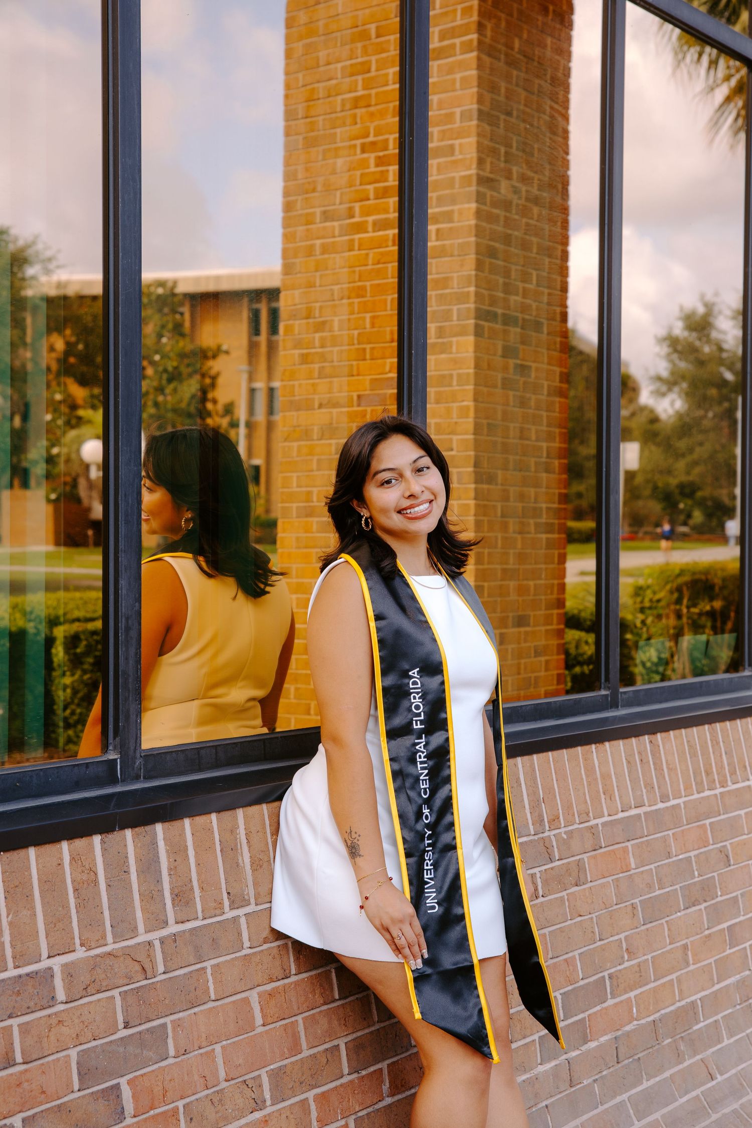 Graduate in white dress with graduation stole poses against brick and glass building.