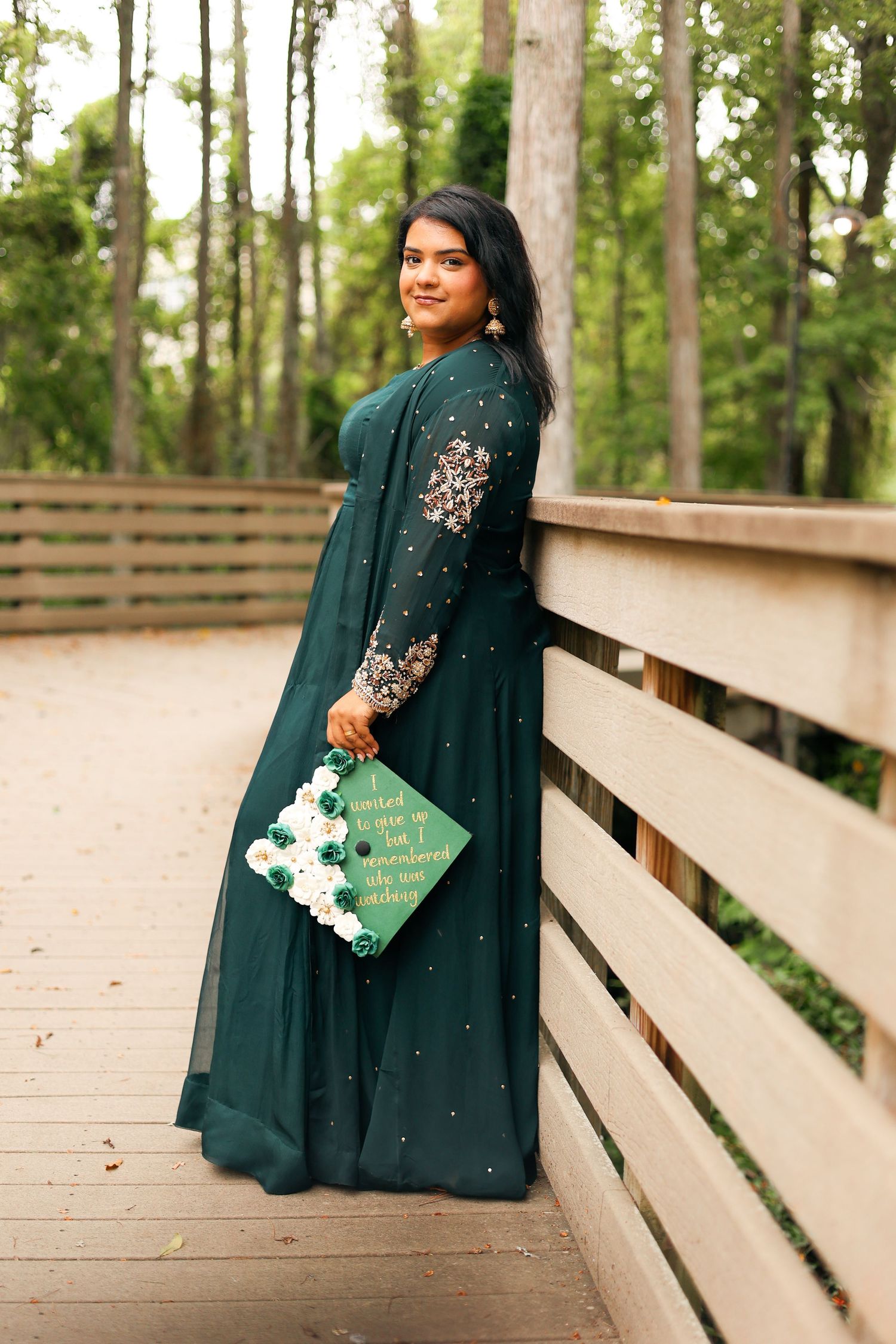 Graduate in emerald green gown poses on wooden walkway during forest photoshoot.