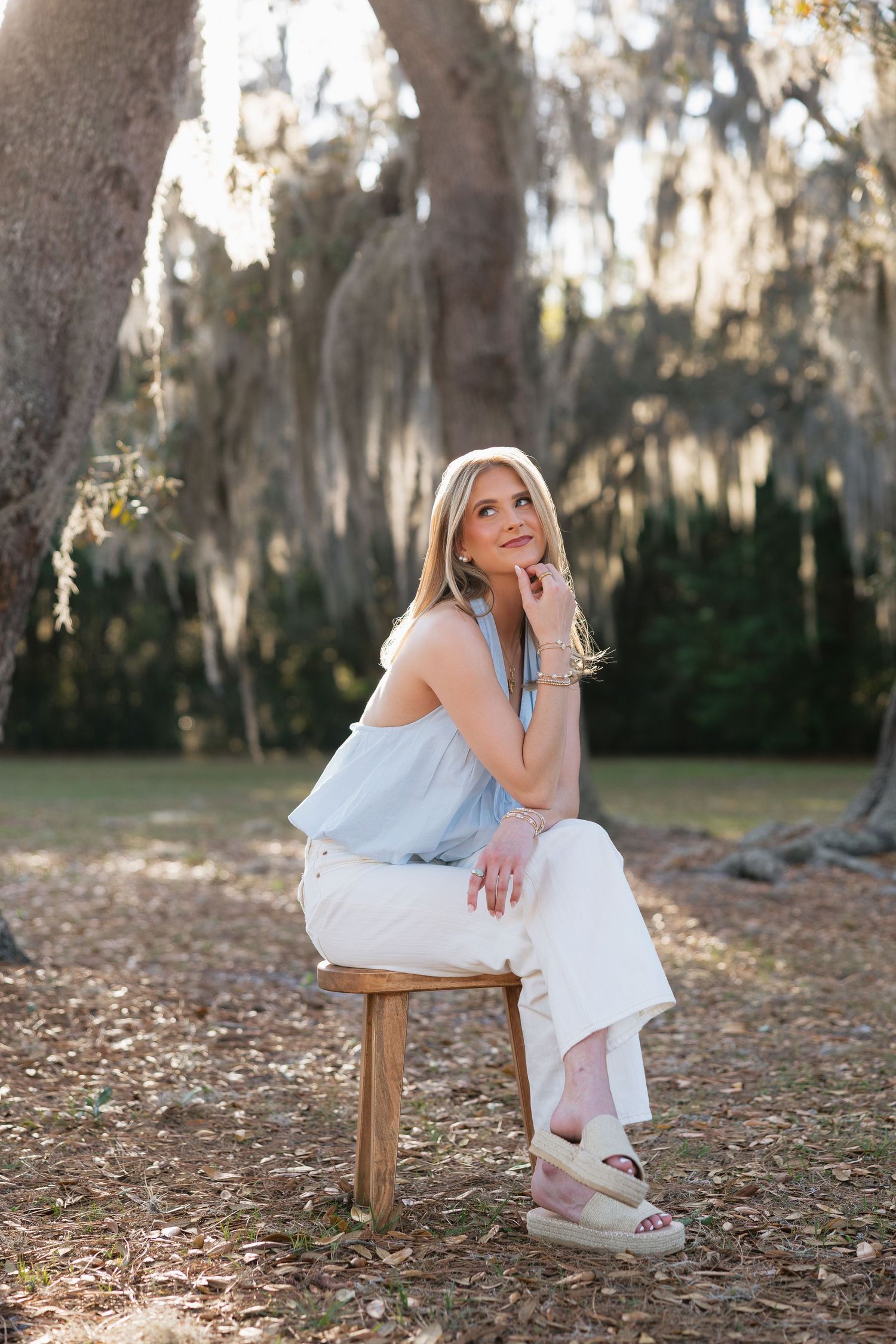 Blonde woman in light blue top and white pants sits on wooden stool outdoors under Spanish moss-draped trees.