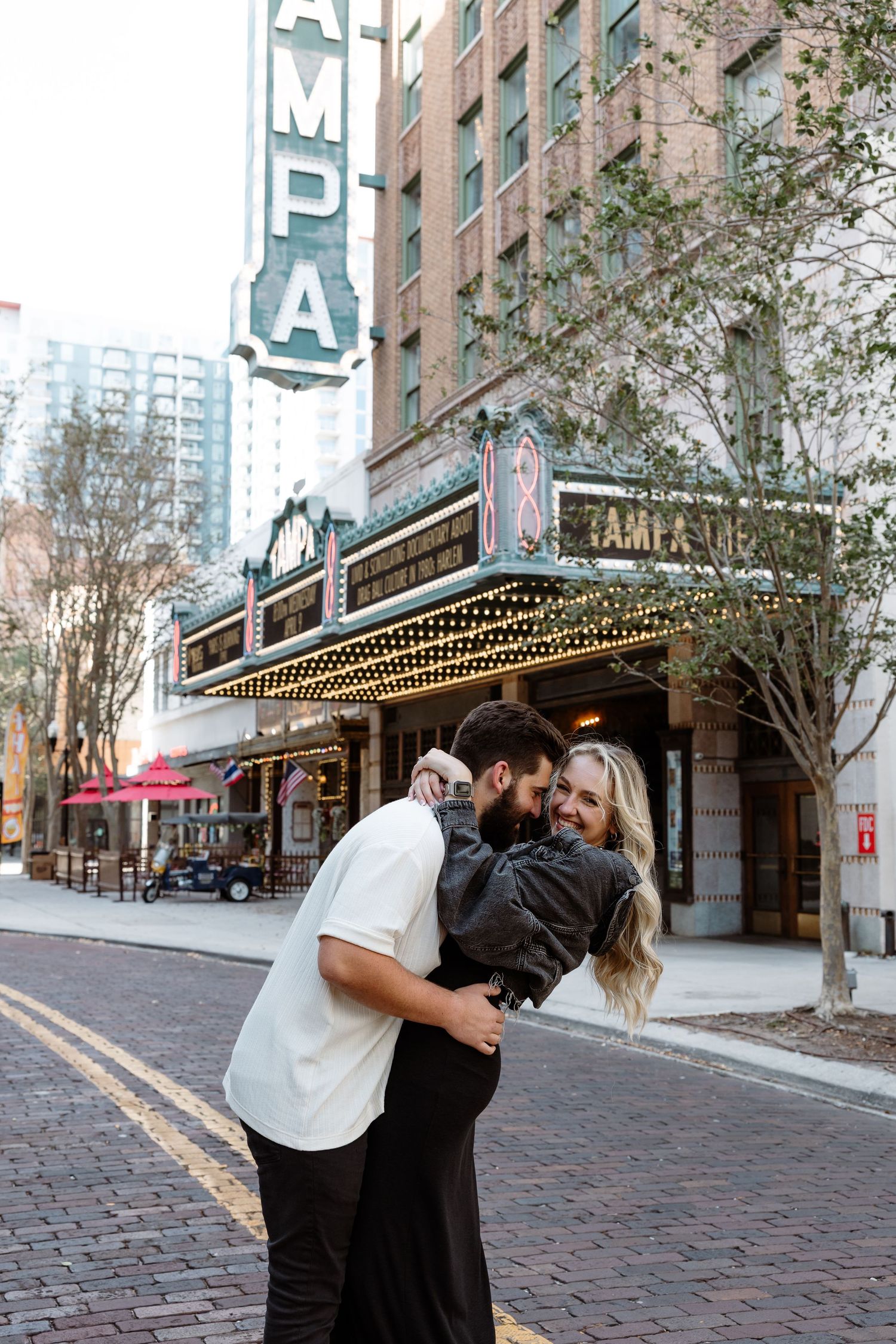 A romantic couple embraces in front of the historic Tampa Theatre with its vintage marquee and brick street.