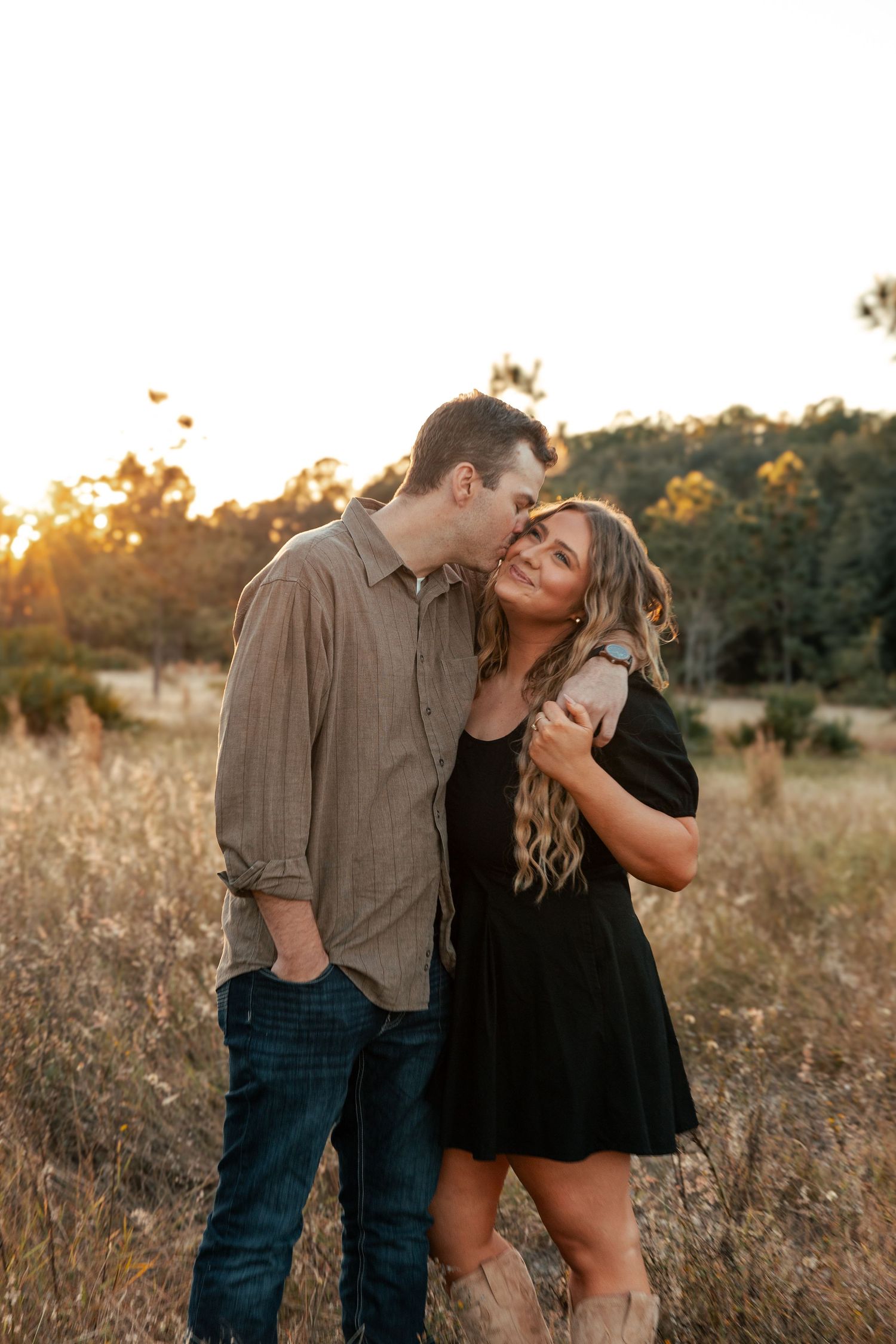A couple shares a tender moment at sunset in a golden field, laughing together in casual attire.