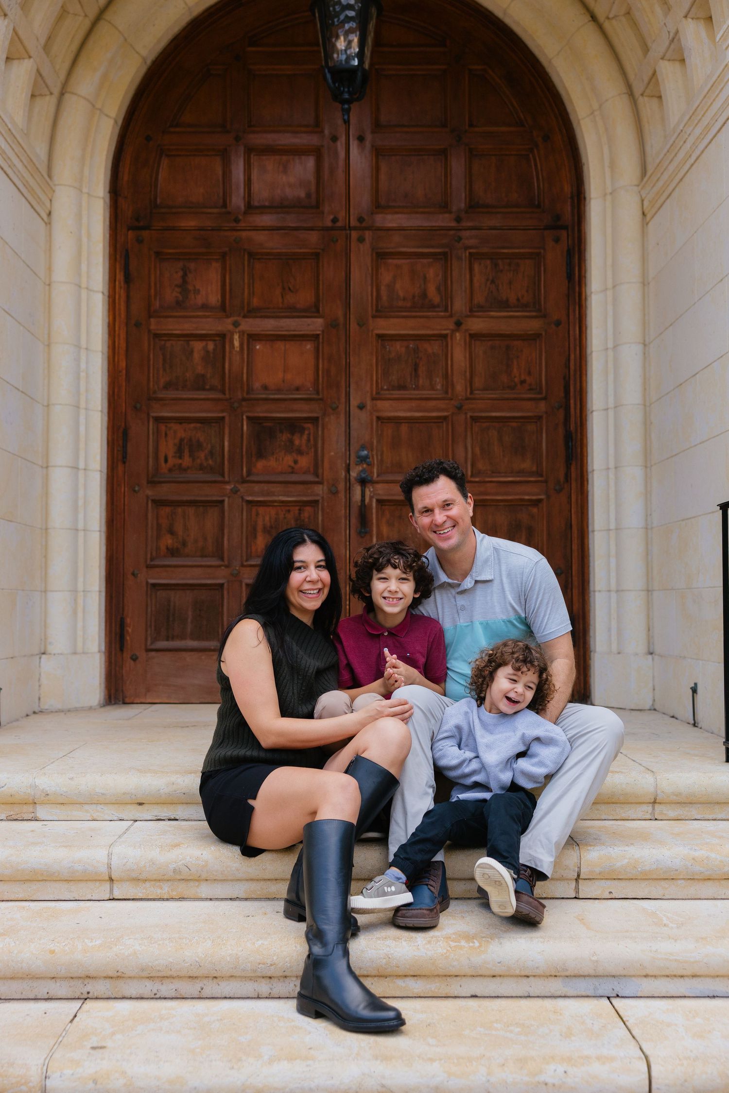 Family sitting and laughing together on stone steps with wooden arched doors in background.
