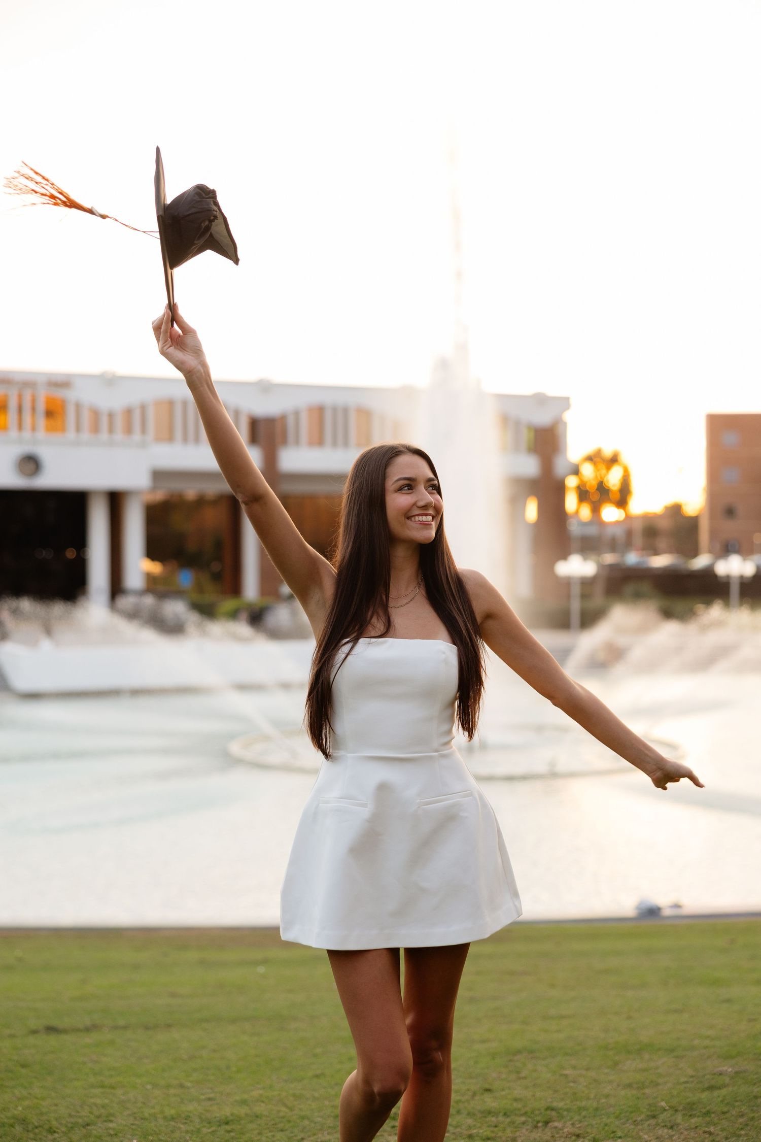 A person in a white dress celebrates outdoors at sunset, raising their graduation cap with joy.