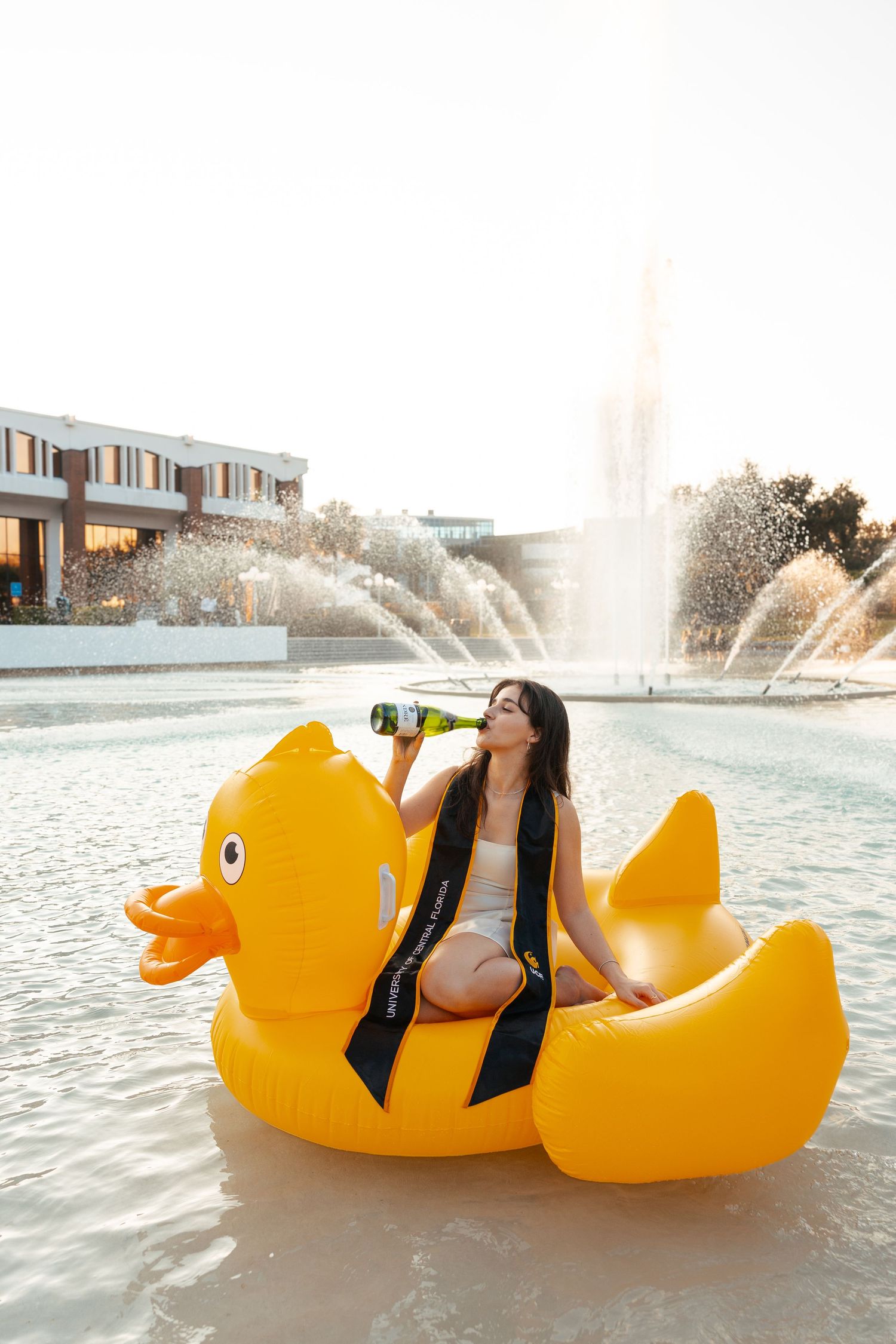 Person relaxing on giant yellow rubber duck float in pool with fountain spraying water in background.