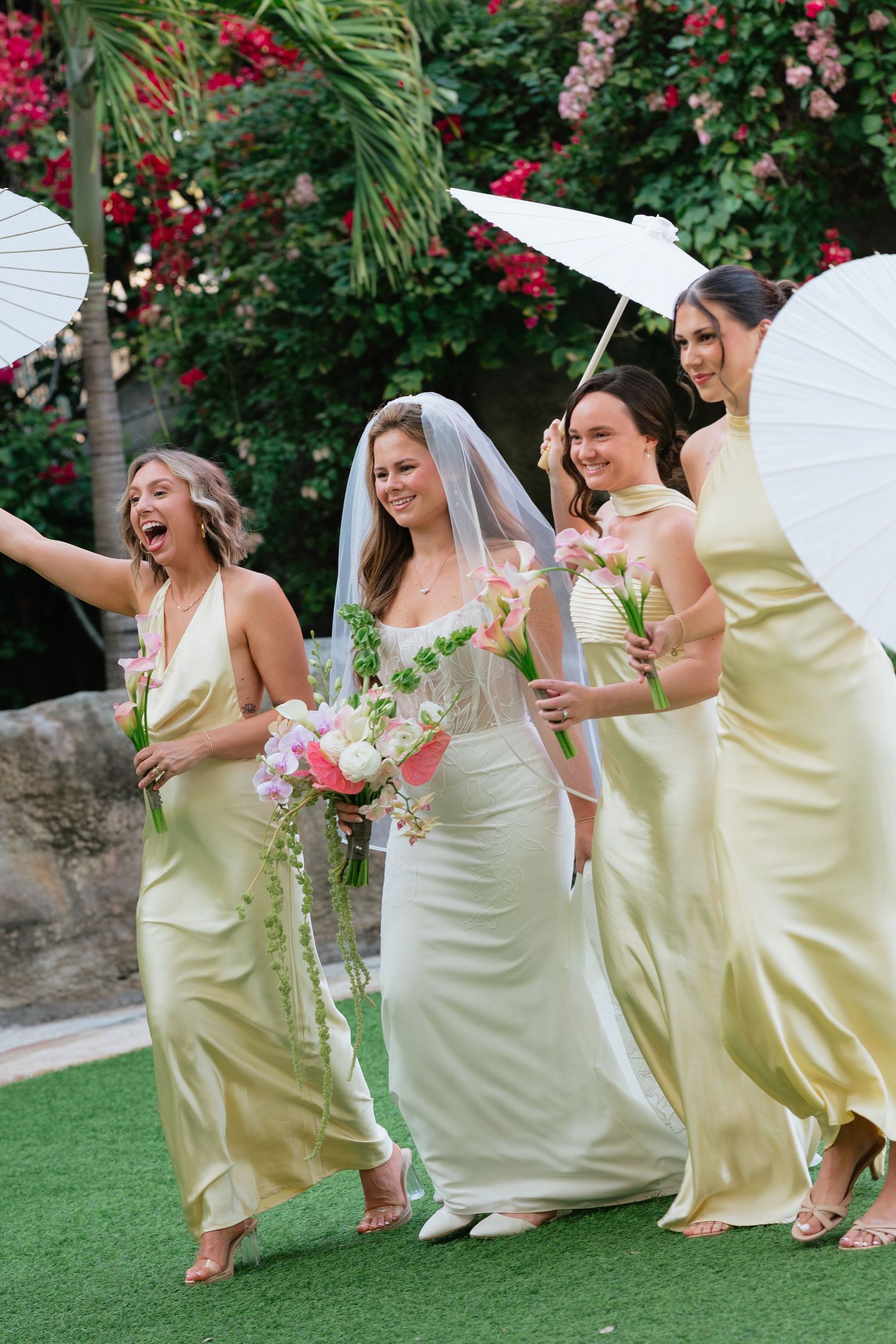 Bridesmaids in pale yellow dresses laughing together on a green lawn with red flowers in background.