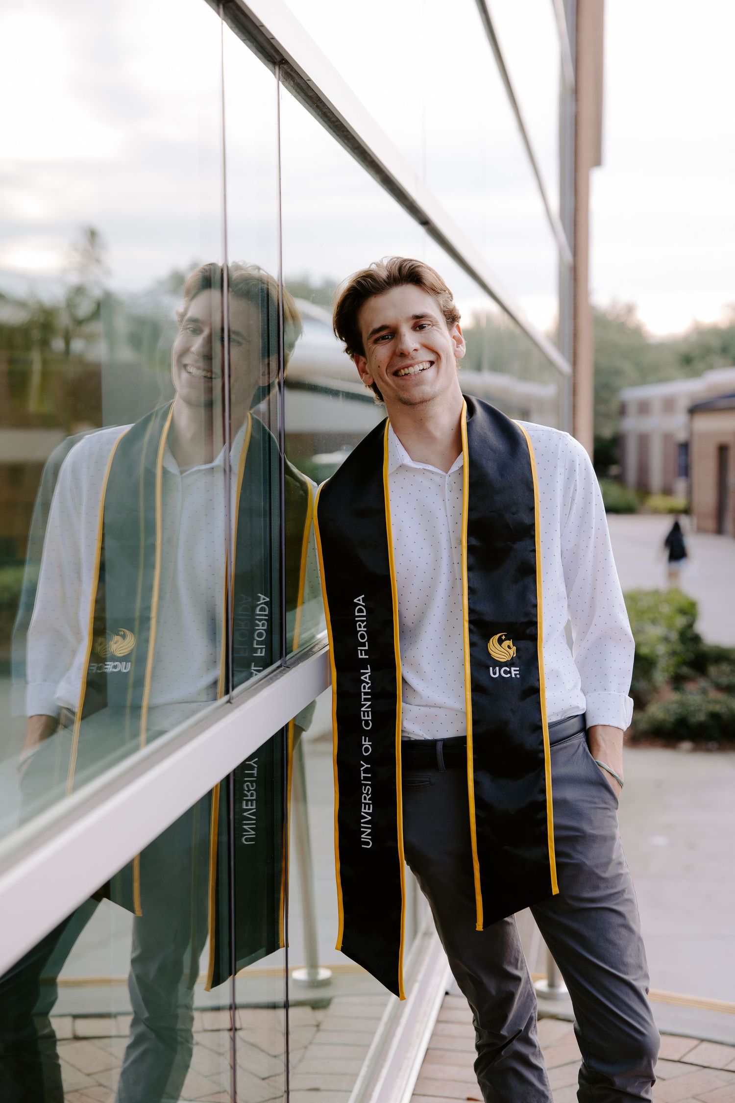 Graduate wearing stole stands against reflective glass building exterior.