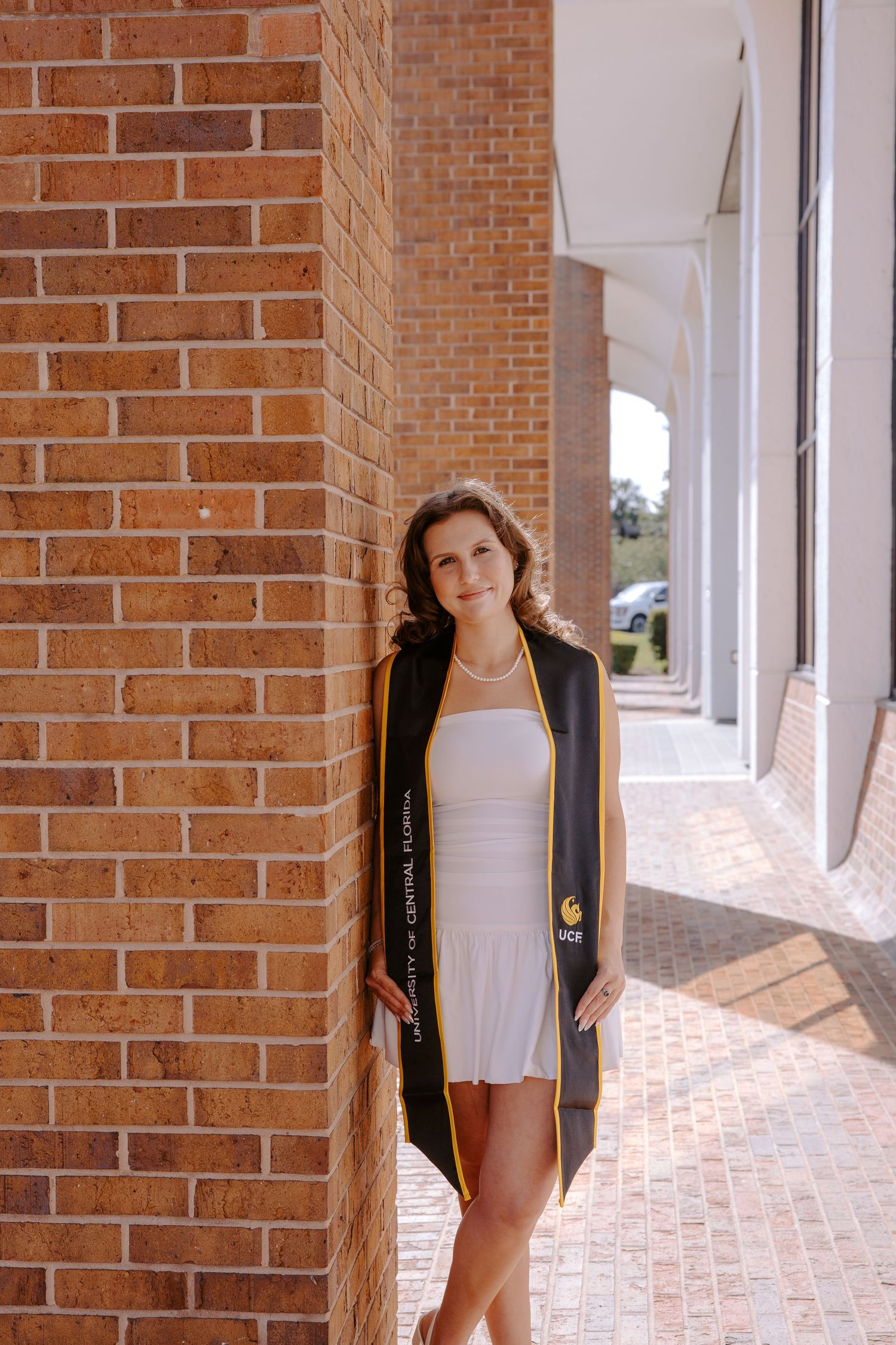 Graduate in black gown poses against brick wall backdrop during sunny photoshoot.