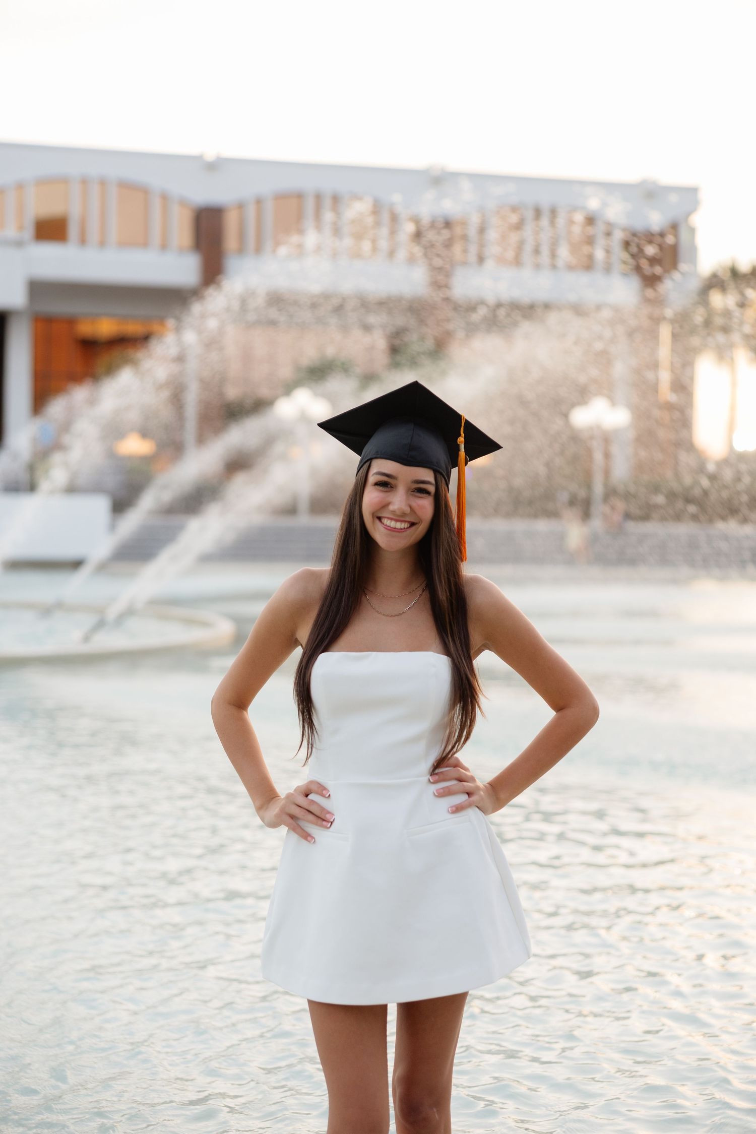 Graduate in white strapless dress and mortarboard cap poses outdoors during sunset.