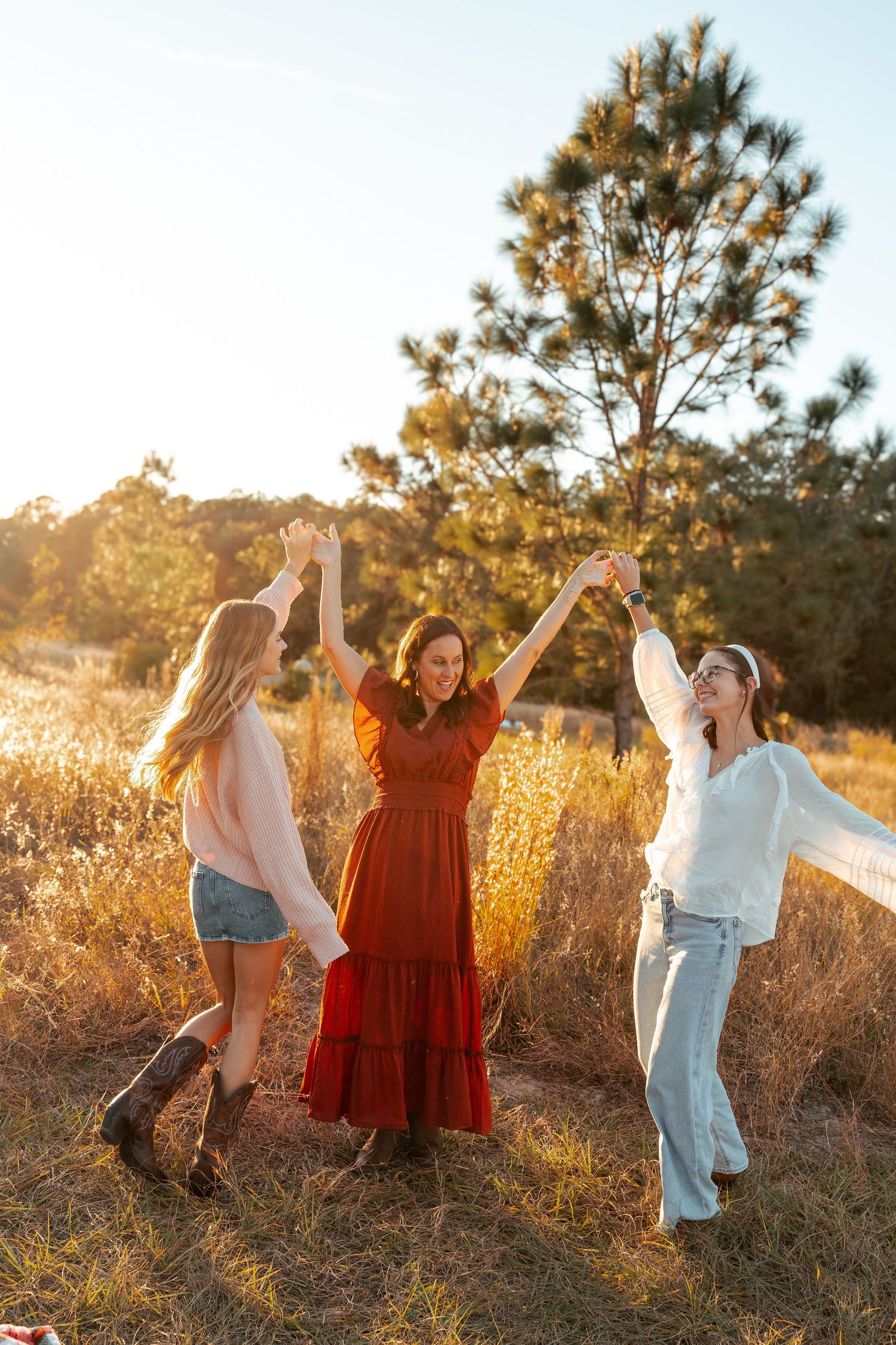 People dance and celebrate joyfully in a golden field at sunset with pine trees in the background.