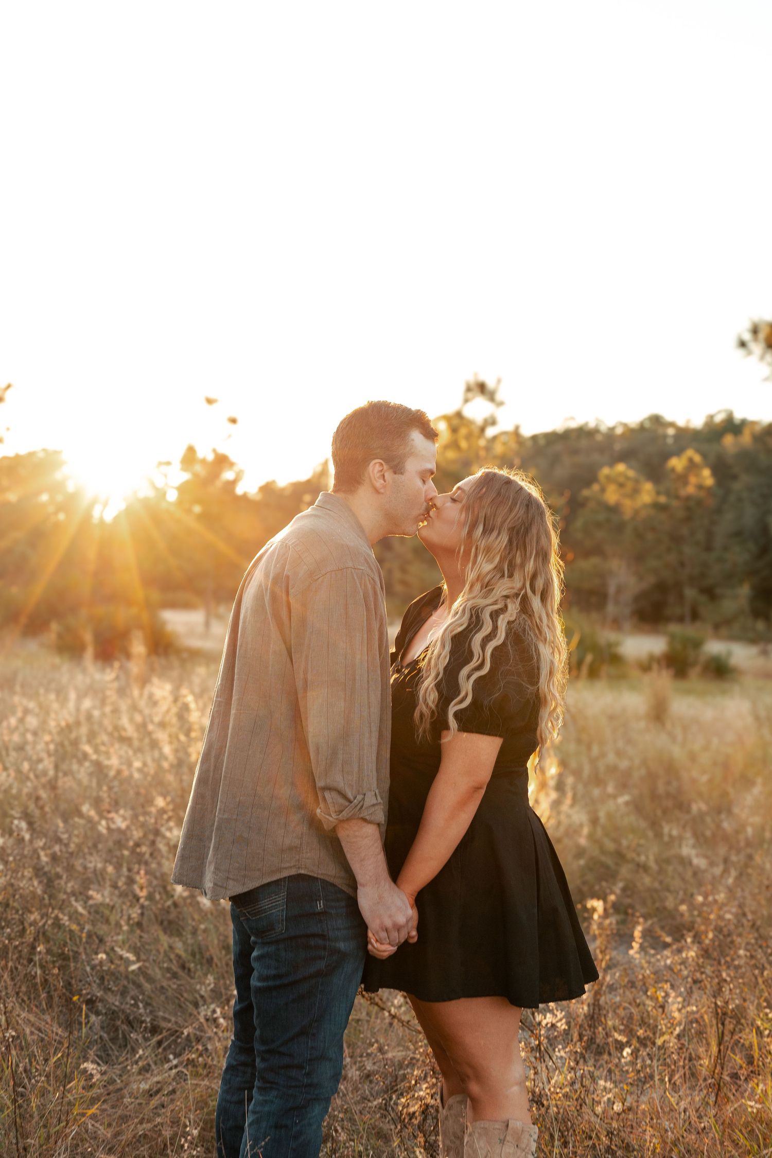 Couple holding hands and kissing at golden sunset in a field of tall dry grass with warm sunlight glowing behind them.