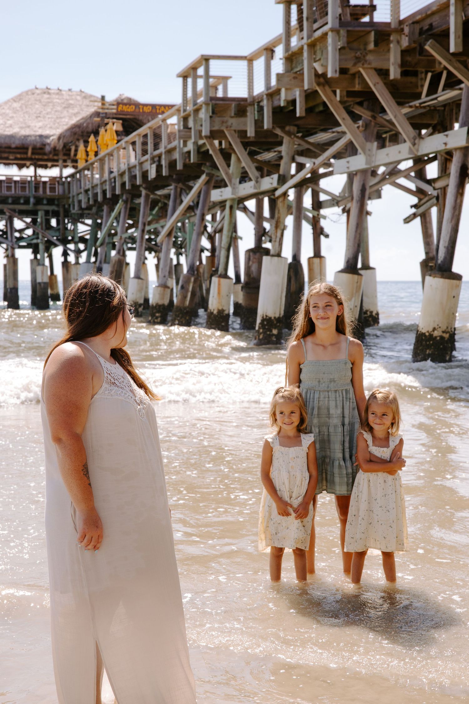 A family wades in shallow water under a wooden pier at the beach.