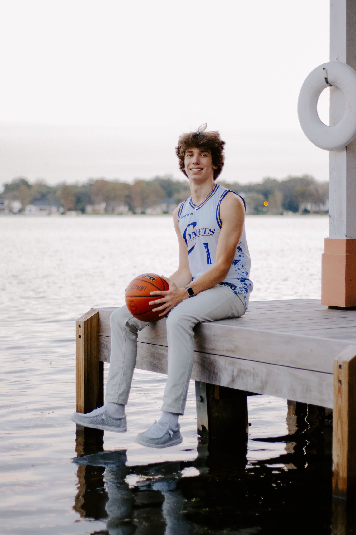 Basketball player sits casually on wooden dock overlooking water.
