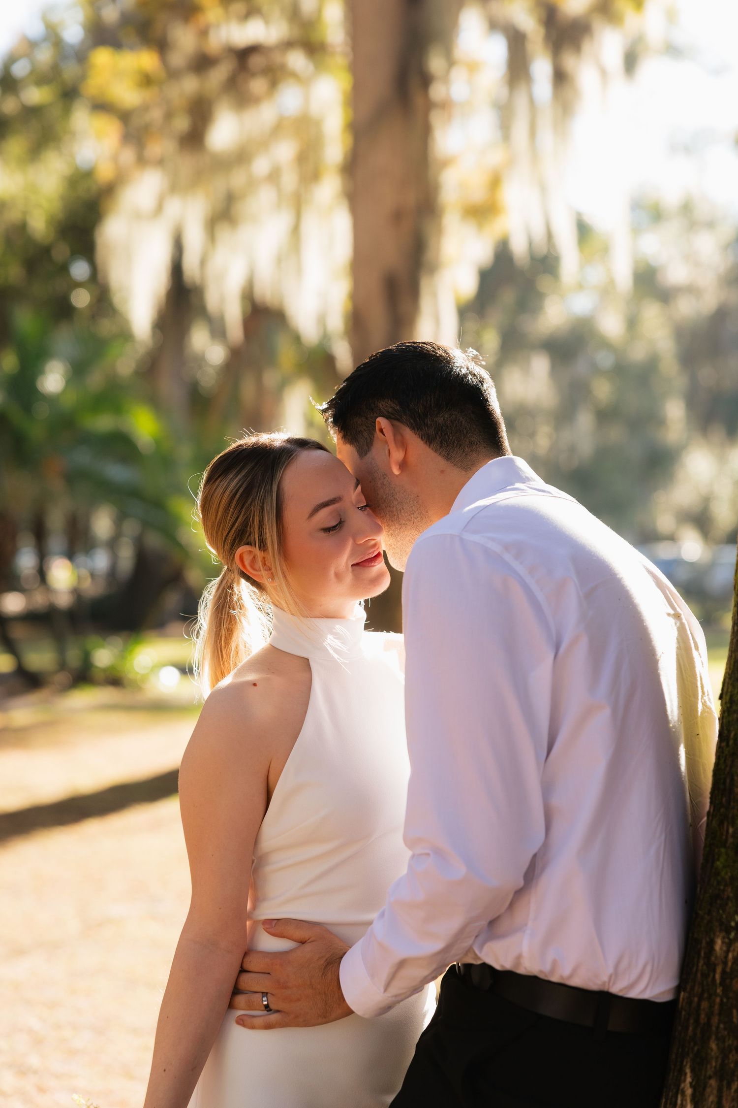 A romantic couple embraces in a sun-dappled garden with Spanish moss hanging from trees in the background.
