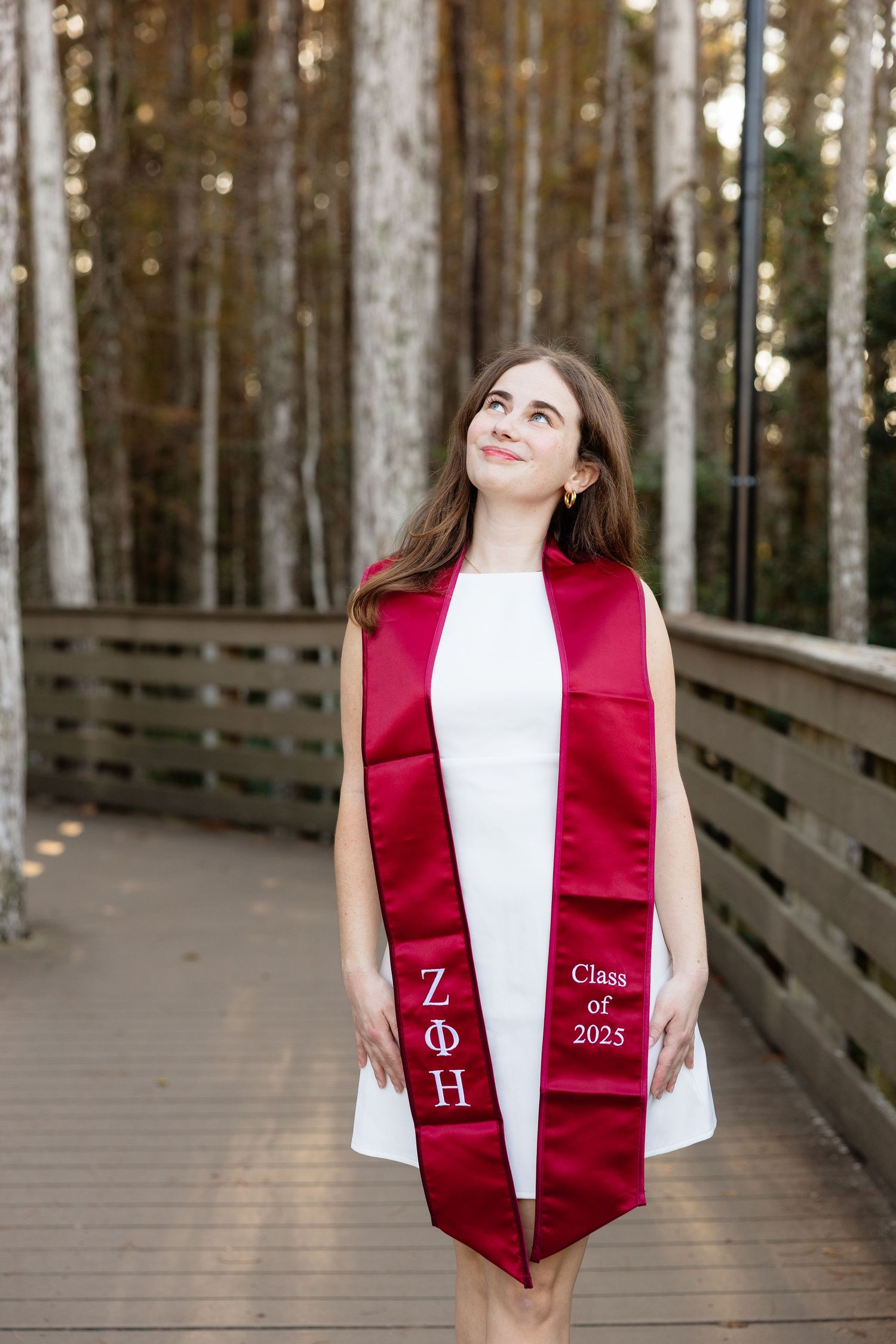 Graduate wearing red and white graduation stole poses on wooden boardwalk surrounded by tall pine trees.