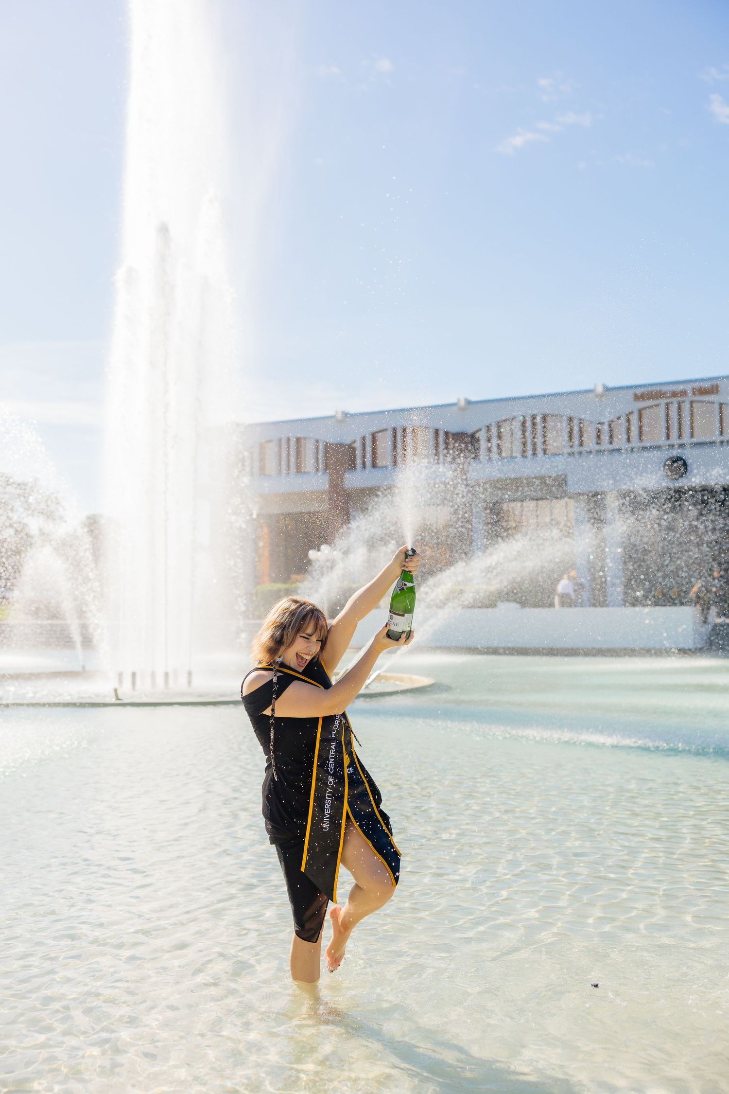 A dancer performs graceful poses in a shallow fountain pool with water sprays and modern buildings in the background.