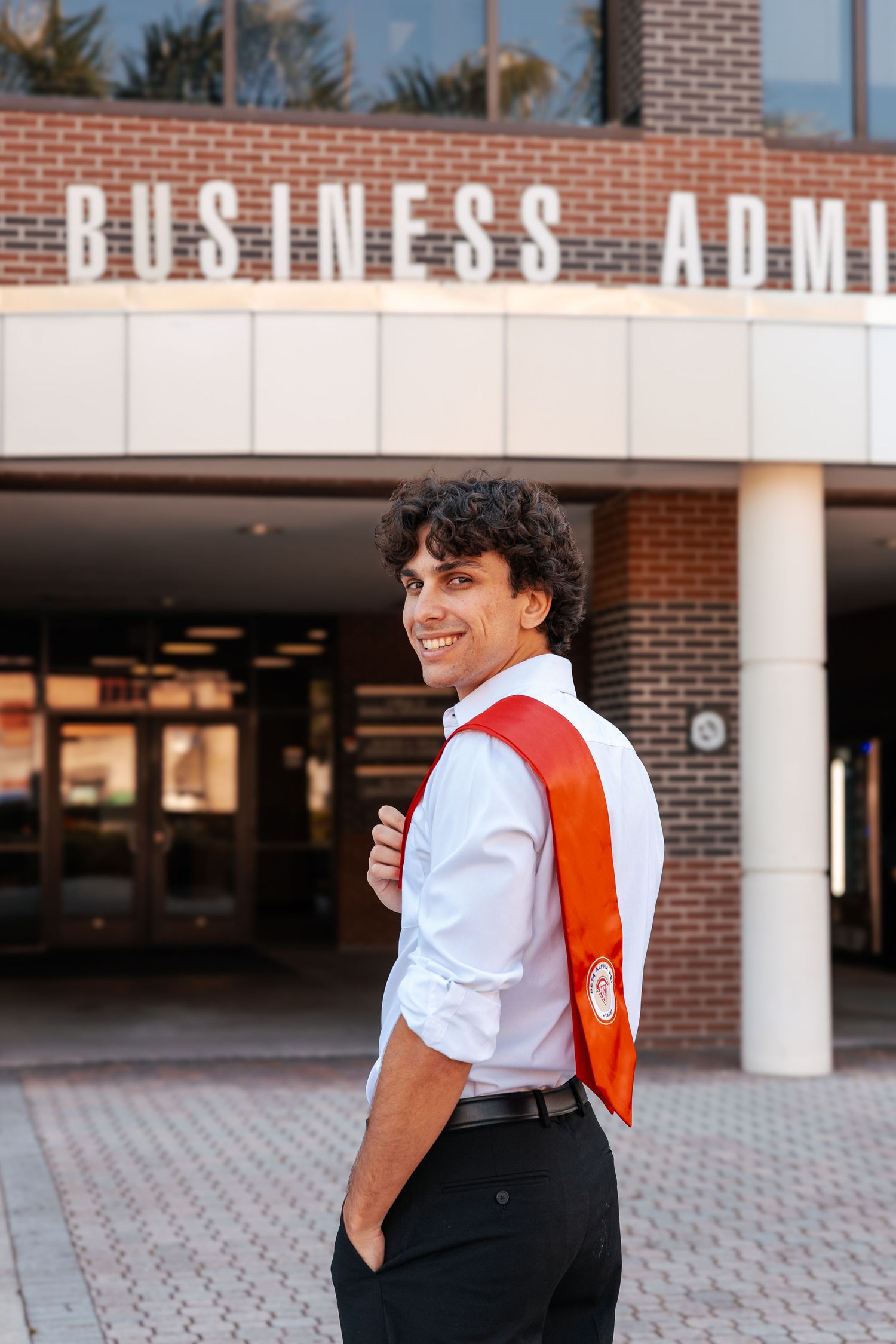 Student wearing red backpack smiles while standing outside business administration building entrance.